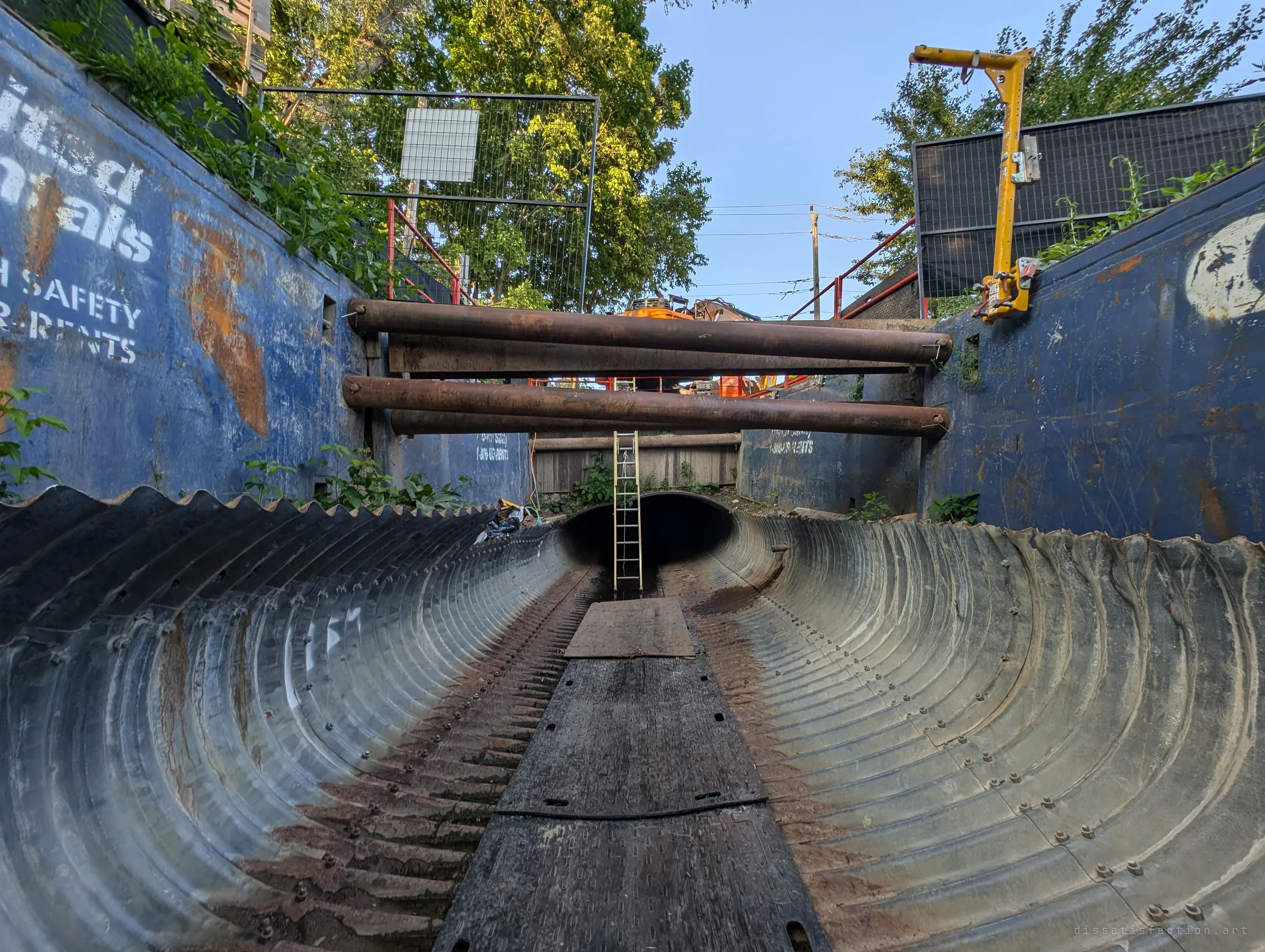 The corrugated pipe lies split open at the bottom of a shored excavation, blue trench boxes and rusted bracing pipes framing a summer sky and trees. A ladder leads up out of the drain.