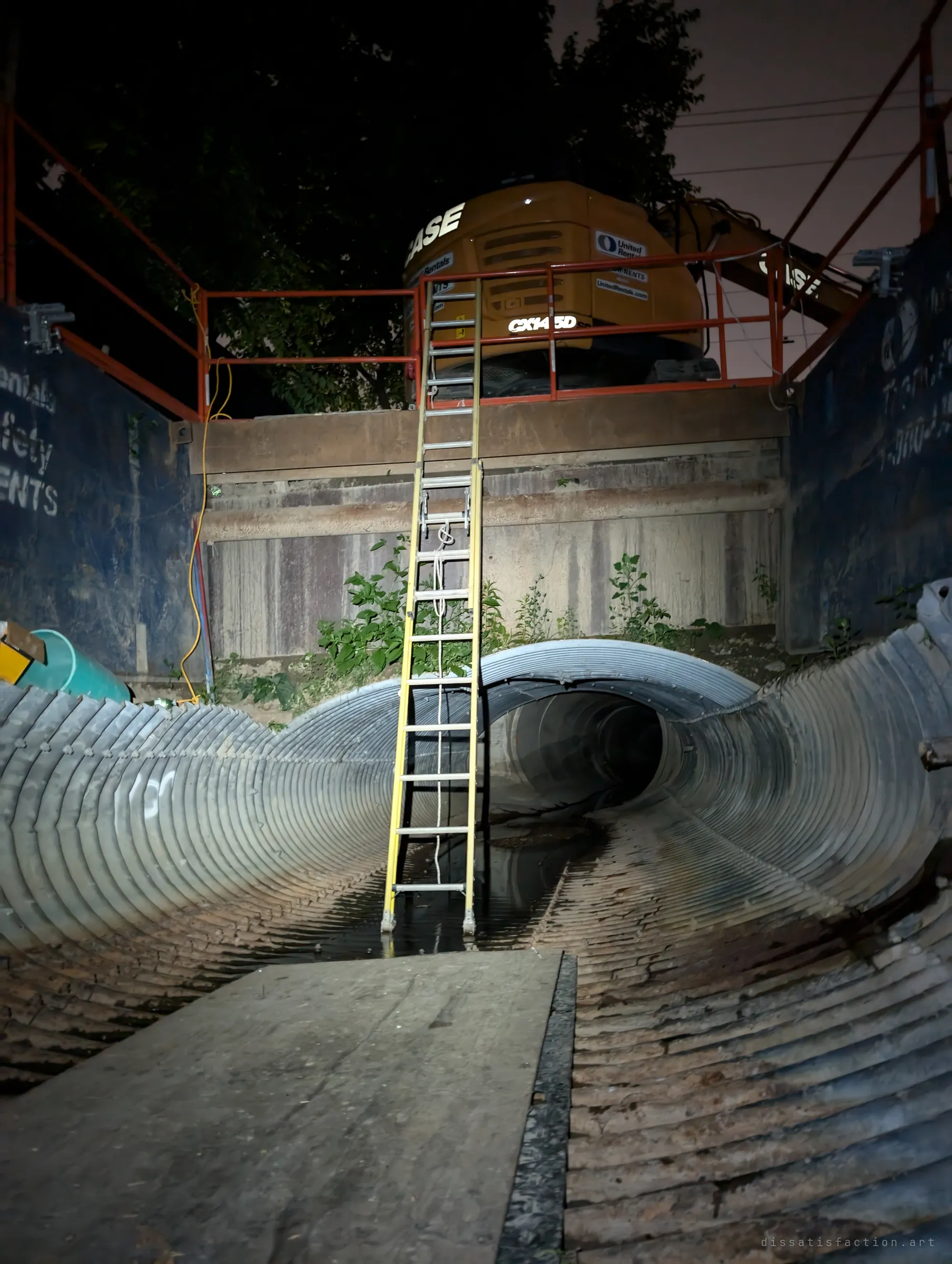 At night, a ladder climbs from the open drain up to an excavator parked behind orange guardrails at the edge of the shaft. The torchlit pipe continues into darkness beneath it.