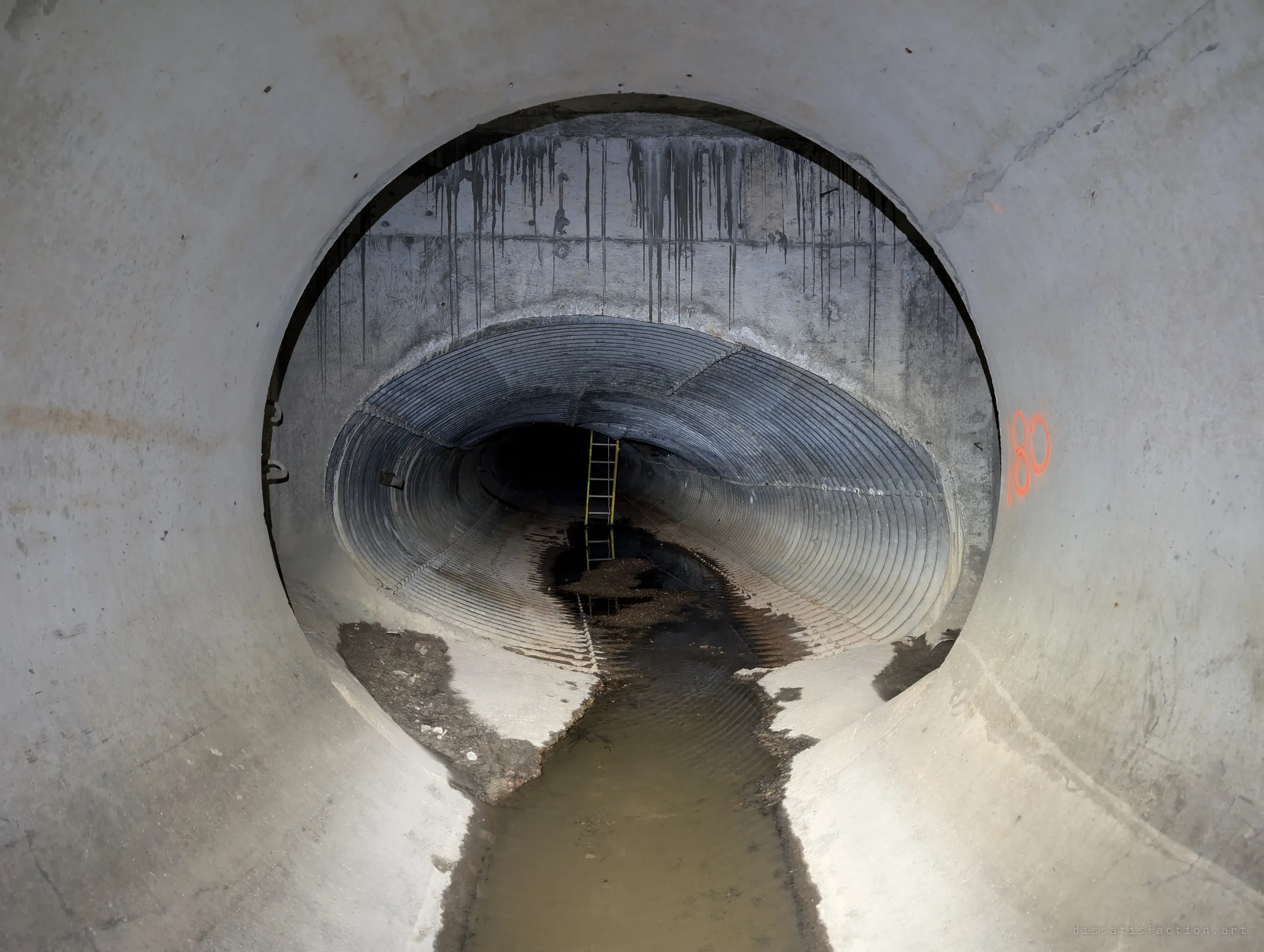 From a round concrete pipe, the view frames a squashed corrugated-steel section beyond, with a yellow ladder standing in the distance and shallow water running down the middle. It marks the change from metal pipe to concrete.
