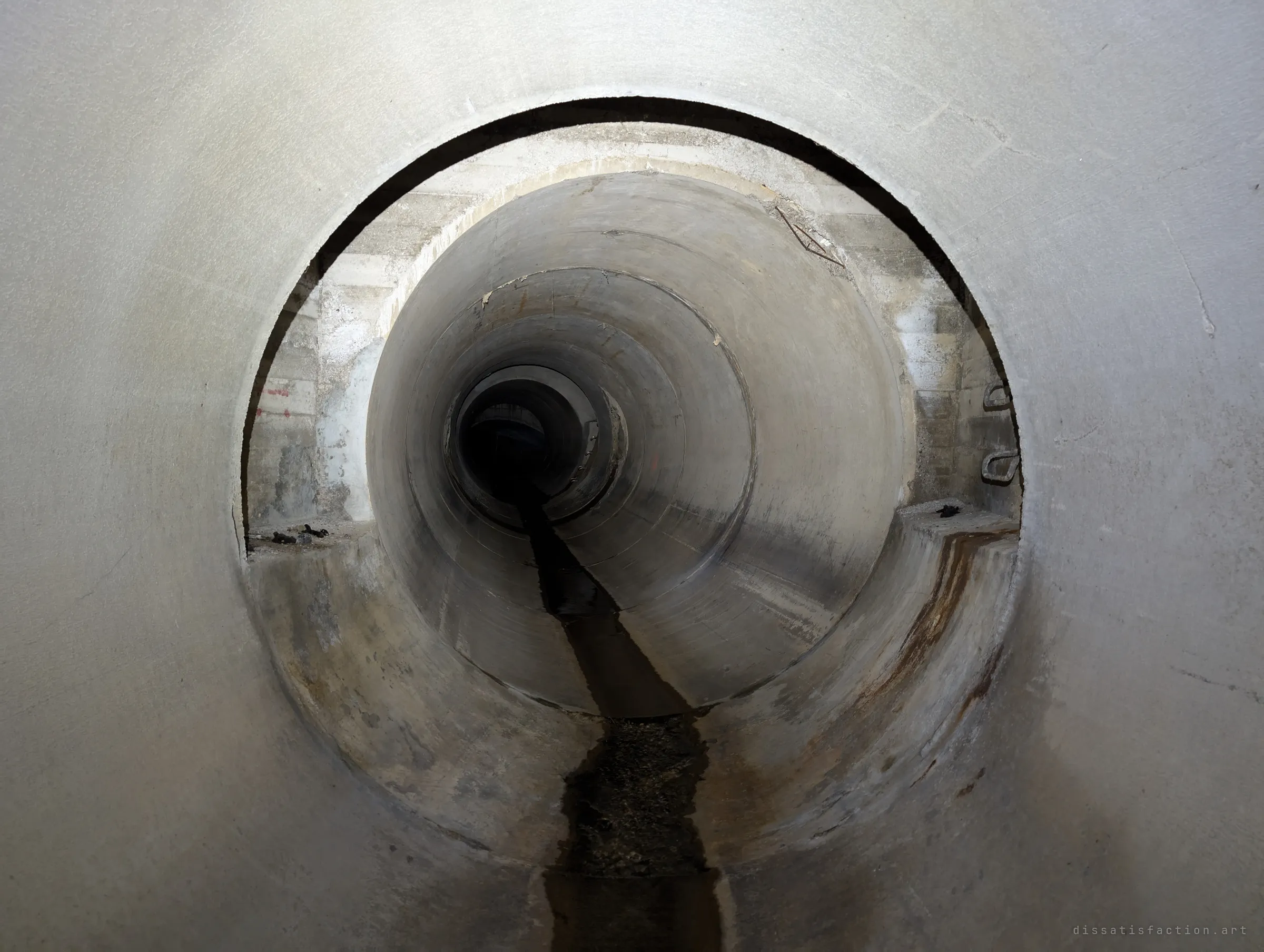 Concentric concrete pipes nest one inside another, a thin dark stream running down the invert into a black vanishing point. The repeating circles pull the eye straight through.