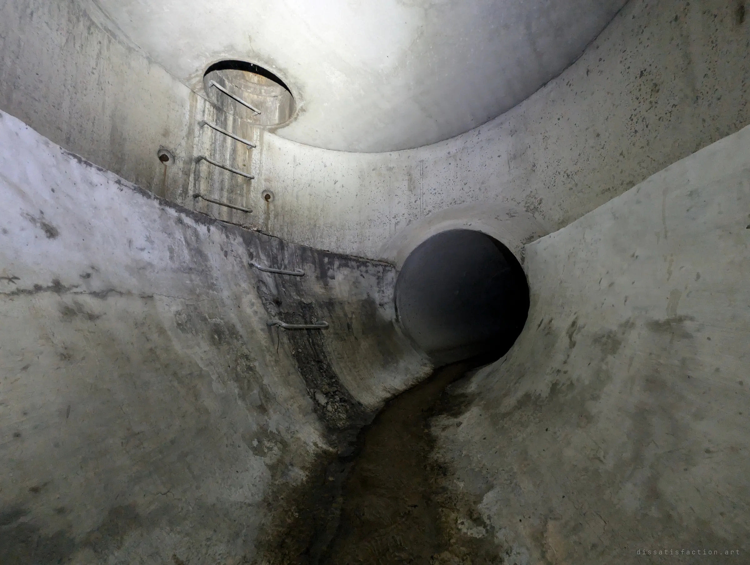 A concrete junction chamber with a domed ceiling, a round access shaft overhead and steel rungs leading up to it. A dark side pipe feeds a muddy channel across the floor.