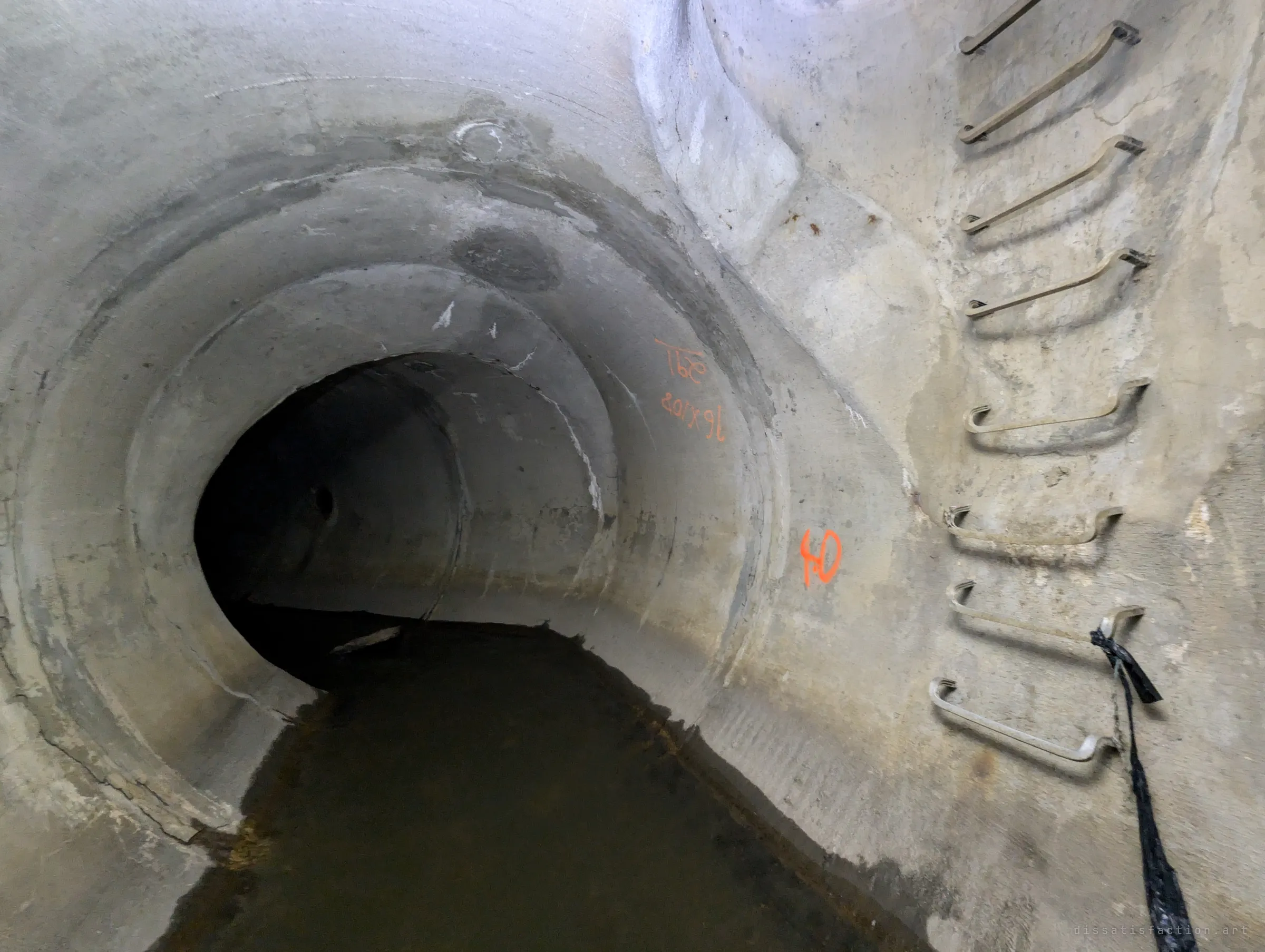A concrete drain bends away to the left under orange survey markings, while a column of steel step irons climbs the wall on the right with a black strap hanging from one rung. Standing water fills the floor.