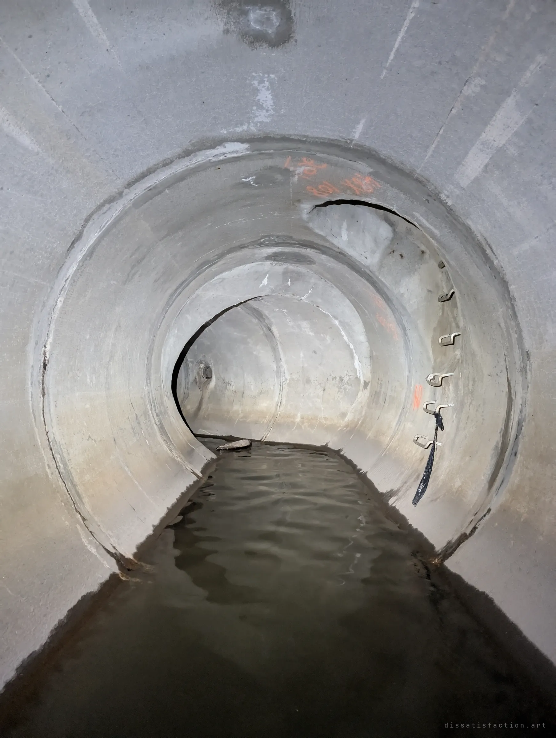 A round concrete pipe stepped down through several ring sections, a thin sheet of water running along its floor under orange survey marks and a column of metal step irons.