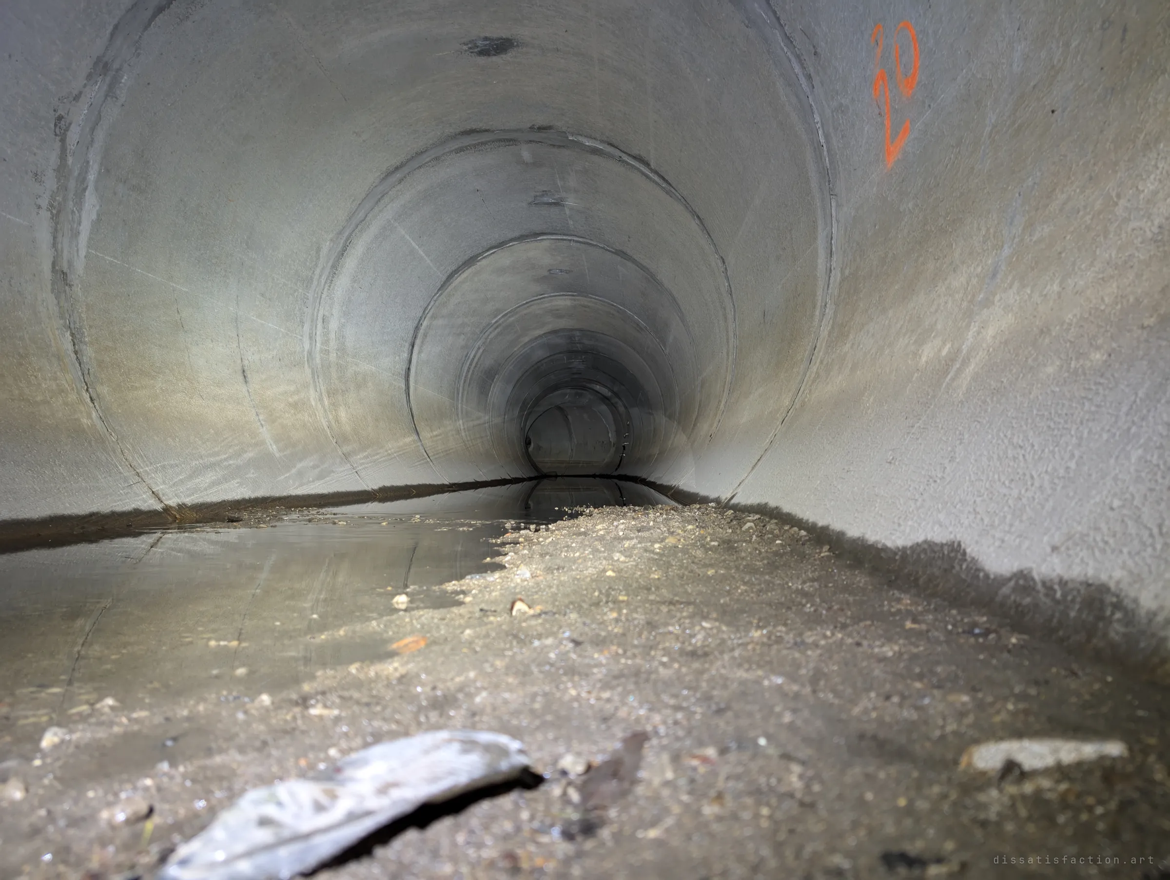 A low view down a long straight concrete pipe, silt and a puddle in the foreground and the joints shrinking into darkness, one orange number sprayed on the wall.