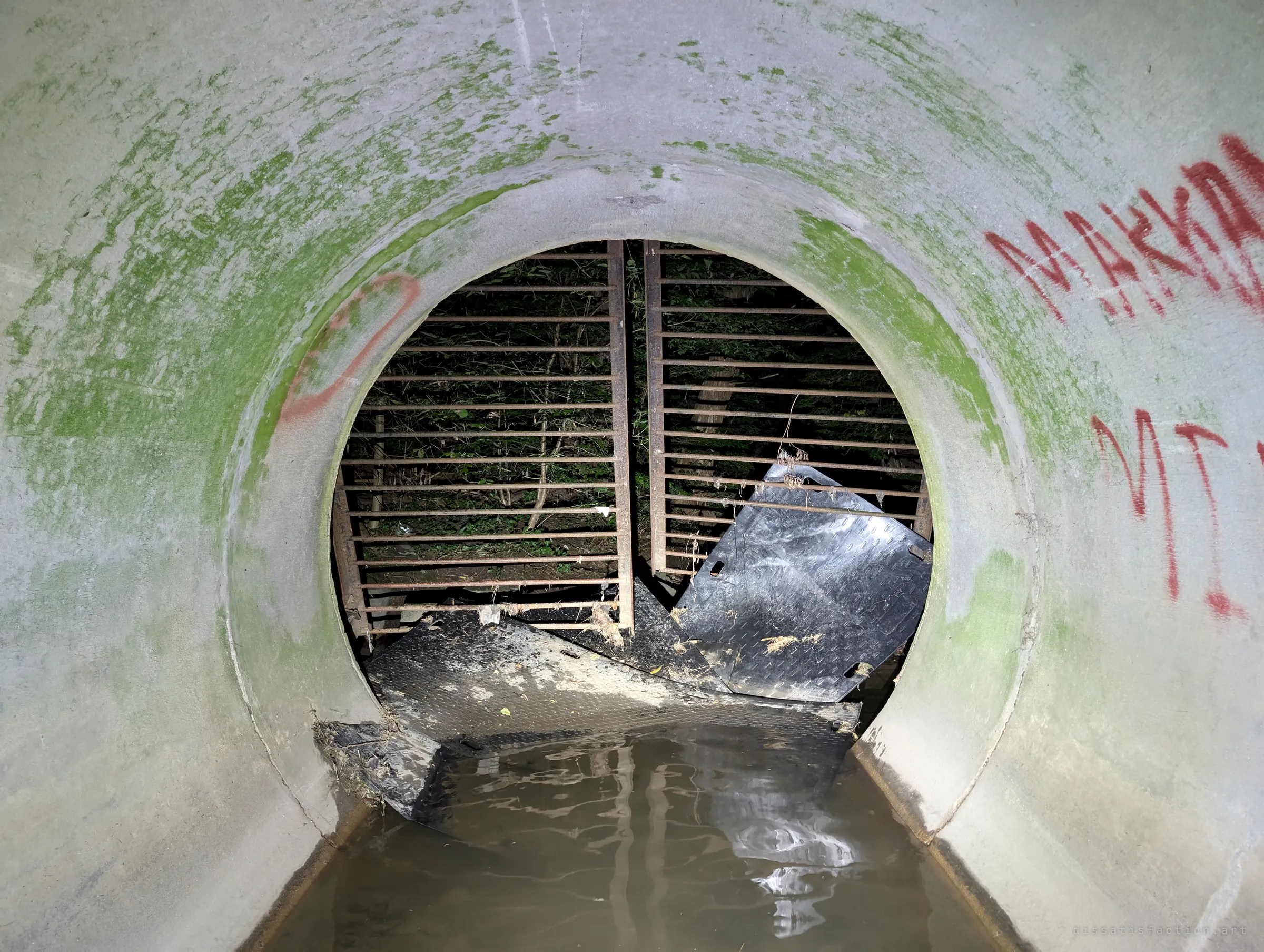 Looking out of a moss-streaked pipe toward the outfall, where a barred gate is bent open and torn steel plates lie in the water, with night foliage beyond.
