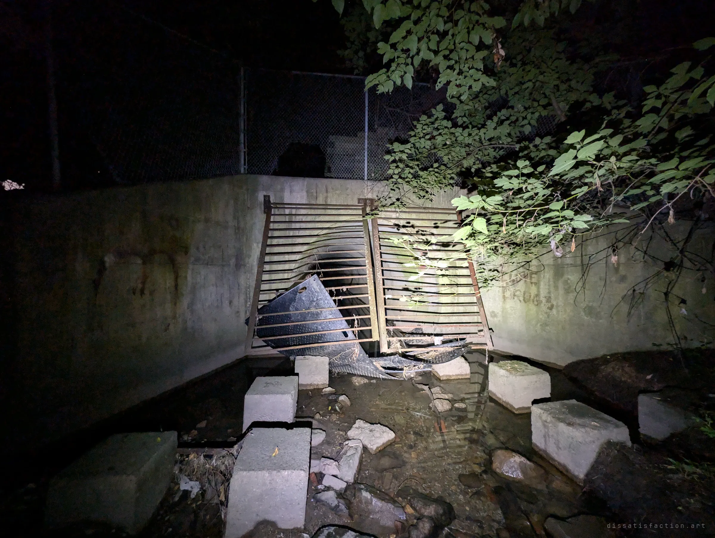 The outfall from outside at night: a concrete headwall with its barred gate forced apart, torn plates caught in it and concrete blocks set across the shallow stream.