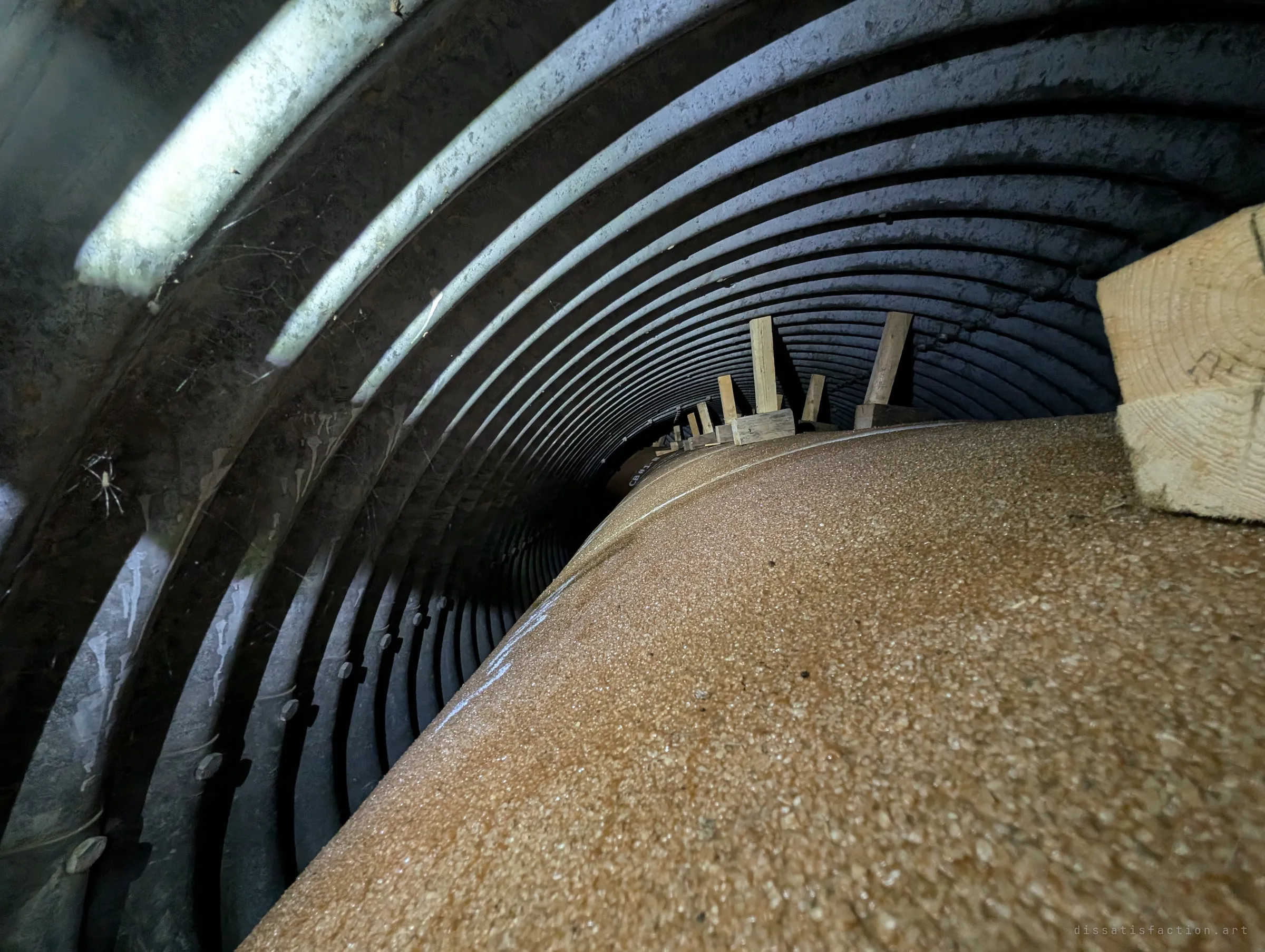 Inside a corrugated steel arch culvert being relined, a gritty liner panel curving up the floor and timber wedges receding along it.