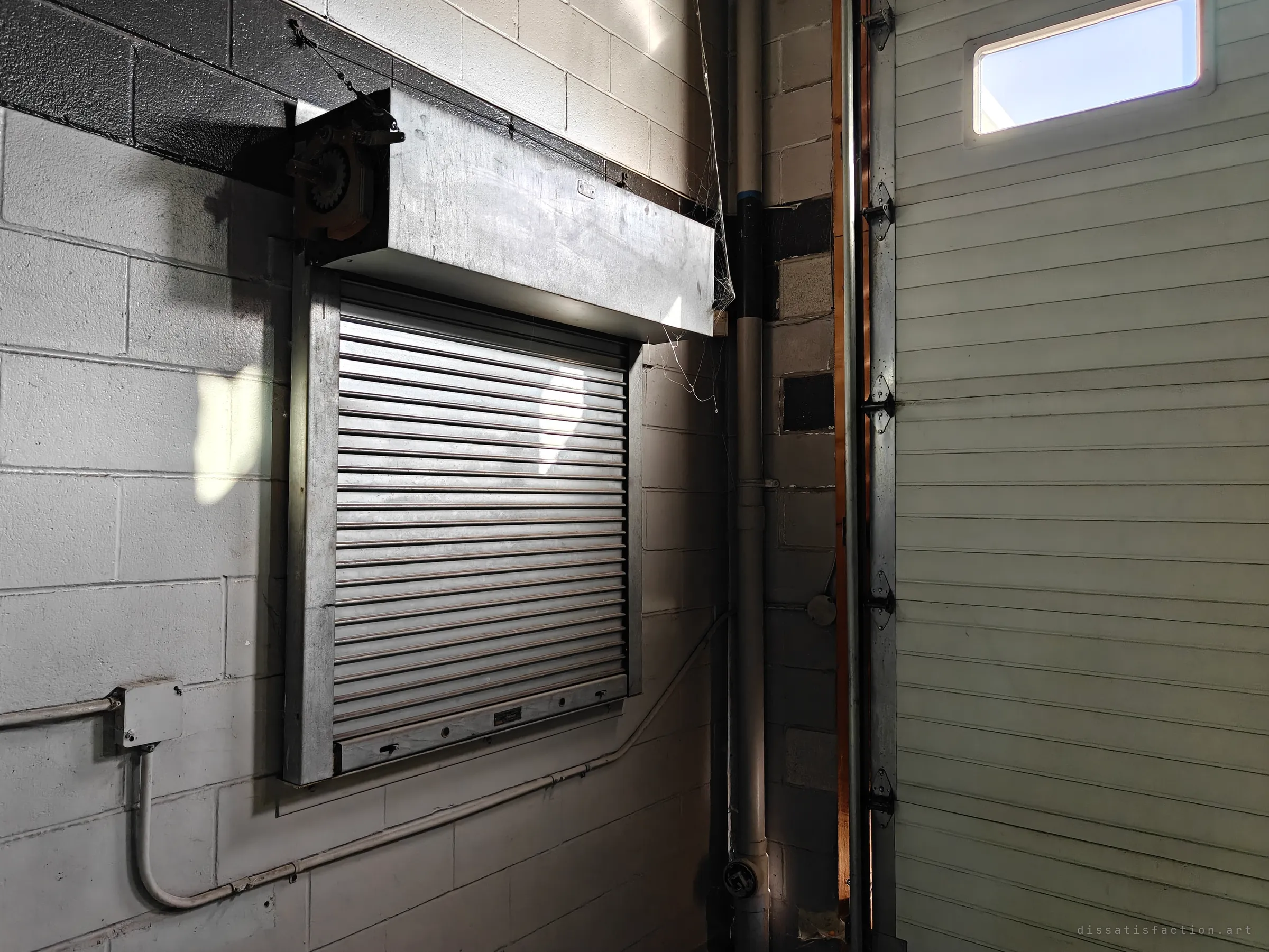 A steel roller shutter and its hood on a white block wall beside a pale sectional door, with conduit and a downpipe running down the corner.