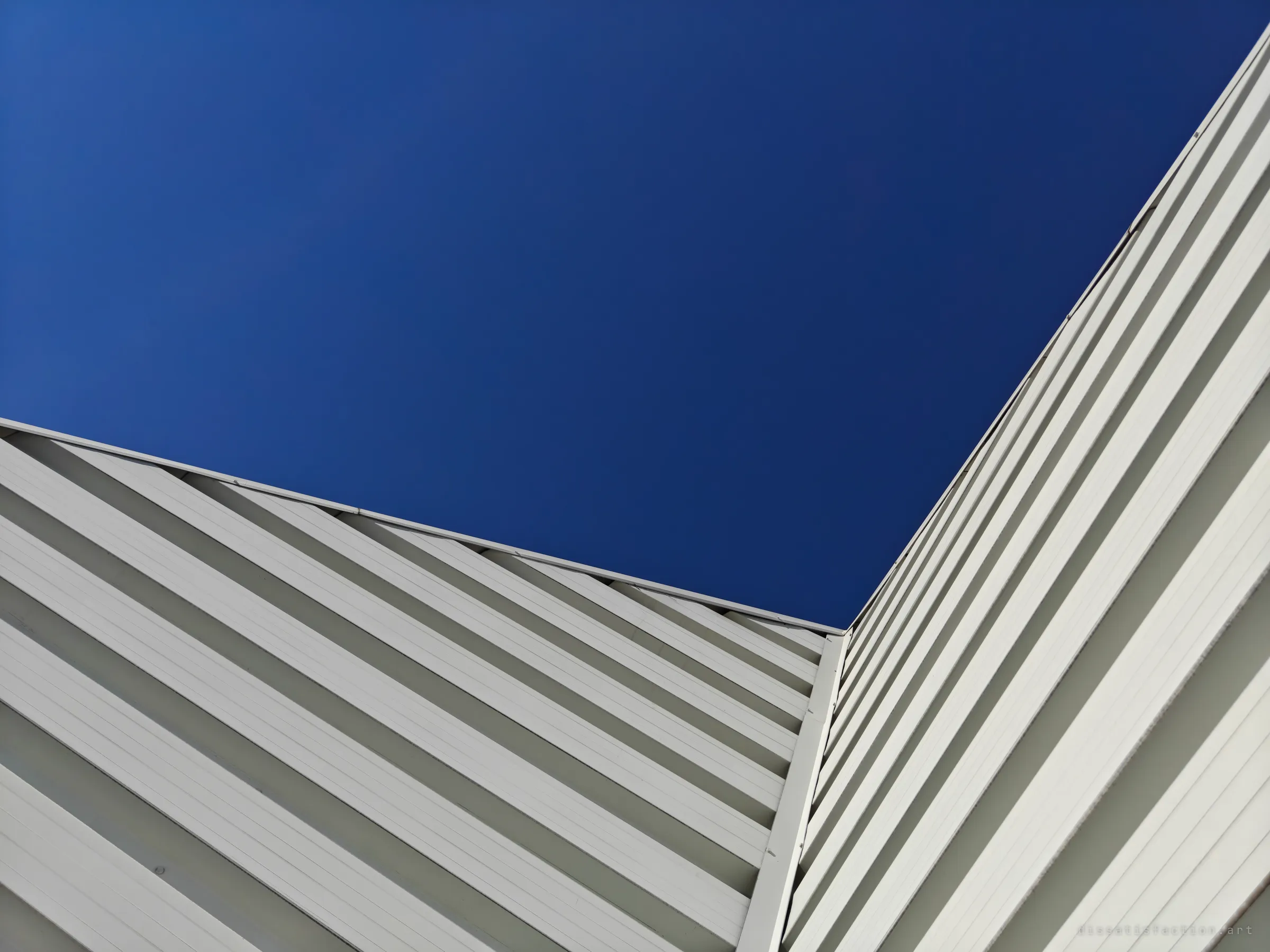 Looking straight up at the corner of a white ribbed metal wall, with its diagonal lines meeting at the roofline against a deep blue sky.