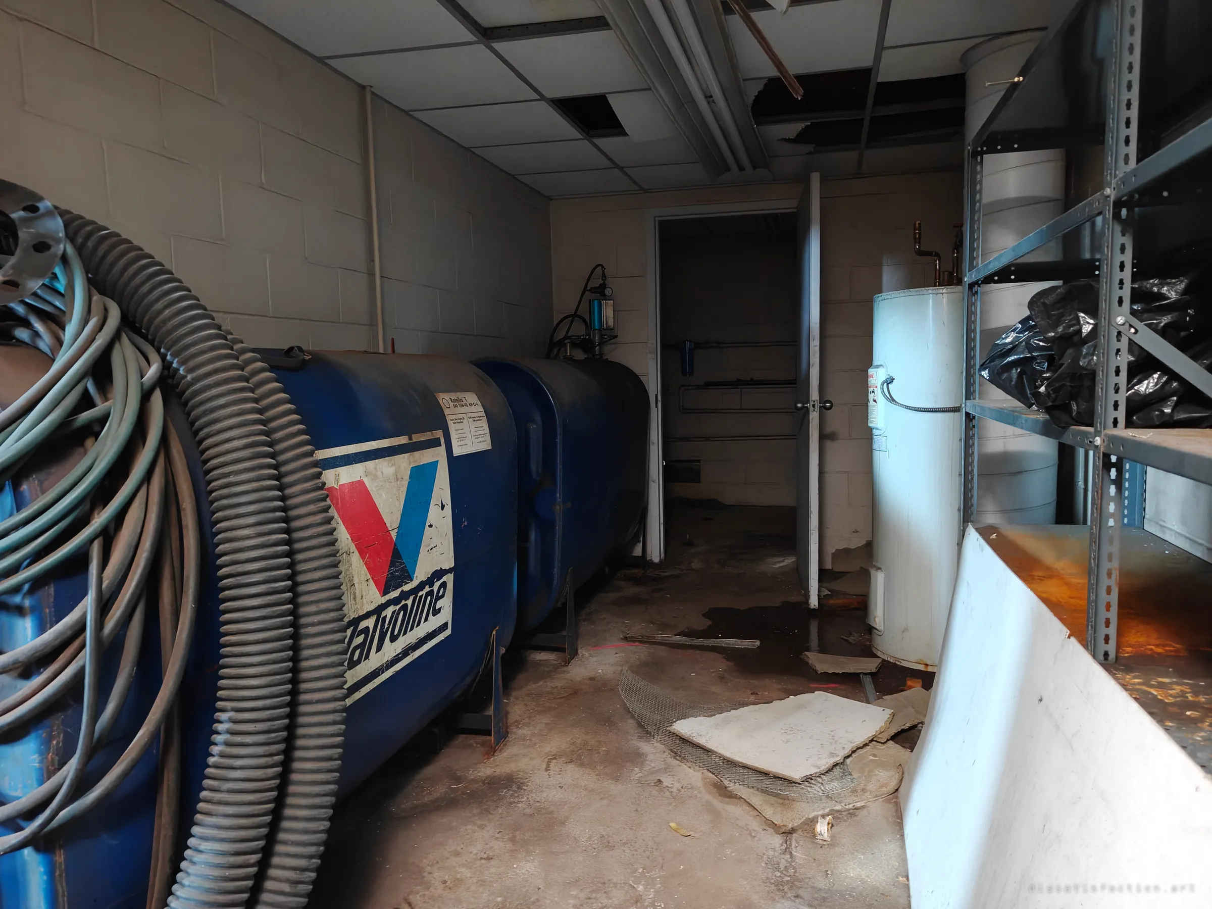 A cramped utility room holds two blue Valvoline oil tanks with coiled hoses, a water heater and steel shelving, with fallen ceiling tiles and stained floor.