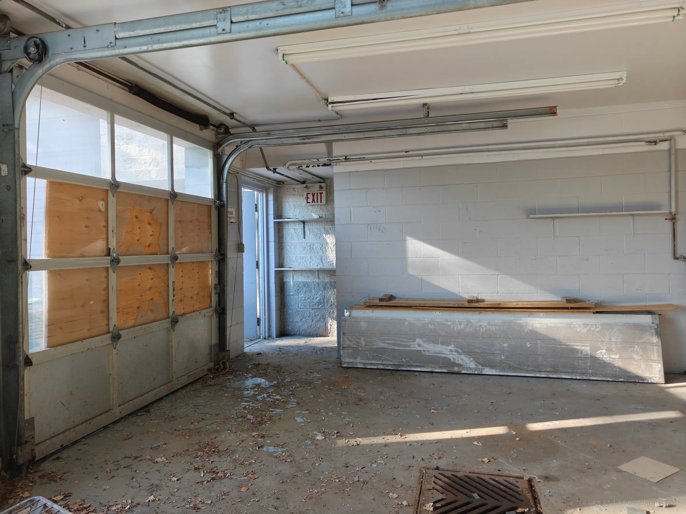 Inside a garage bay, a sectional door with its glass boarded over in plywood, broken glass and dead leaves scattered on the concrete, and a stack of timber leaning on an aluminium panel by the wall.