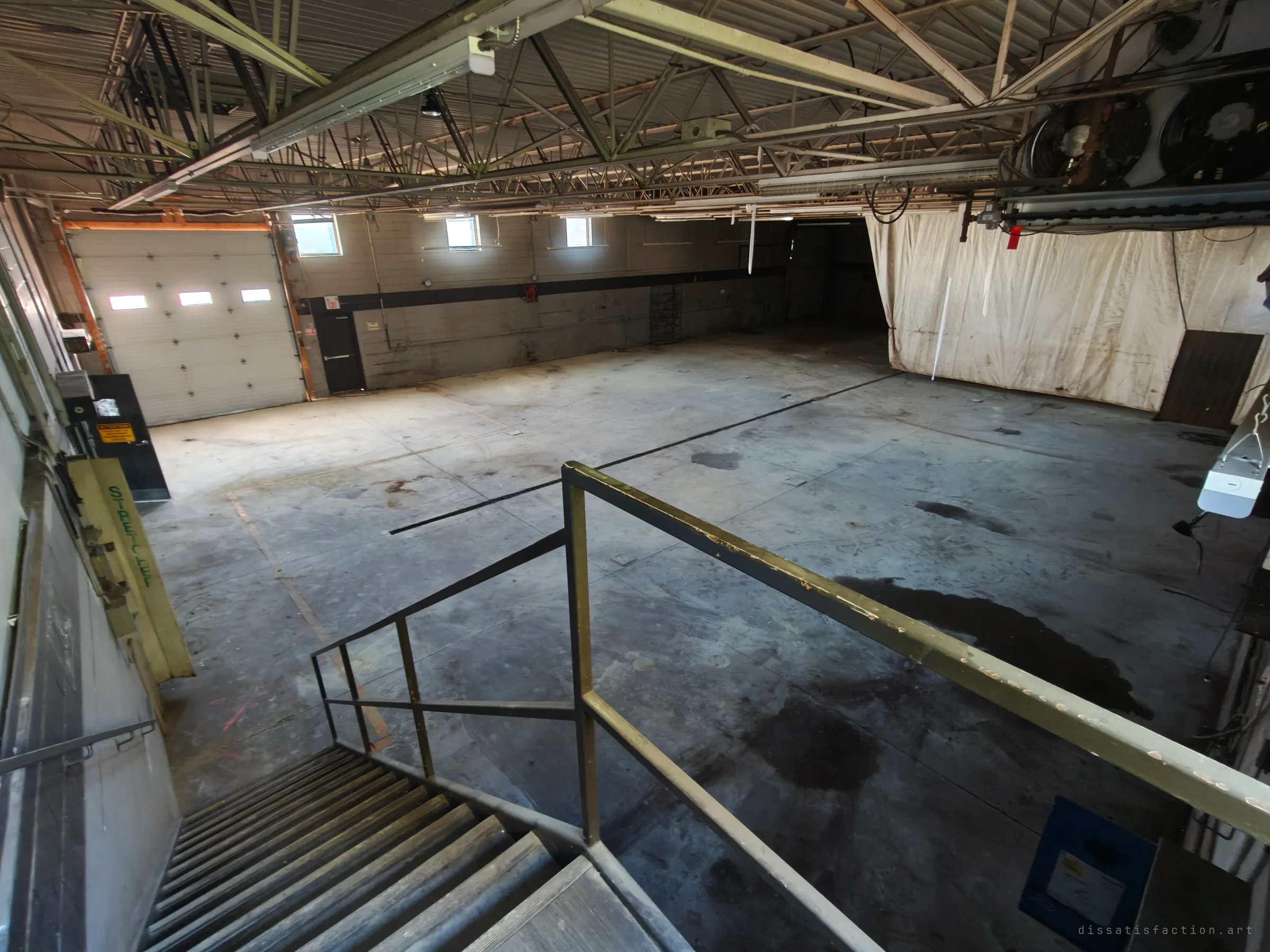 Looking down from a stair landing with a worn metal railing into the empty garage bay, with a roll-up door on the left and a dirty curtain on the right.