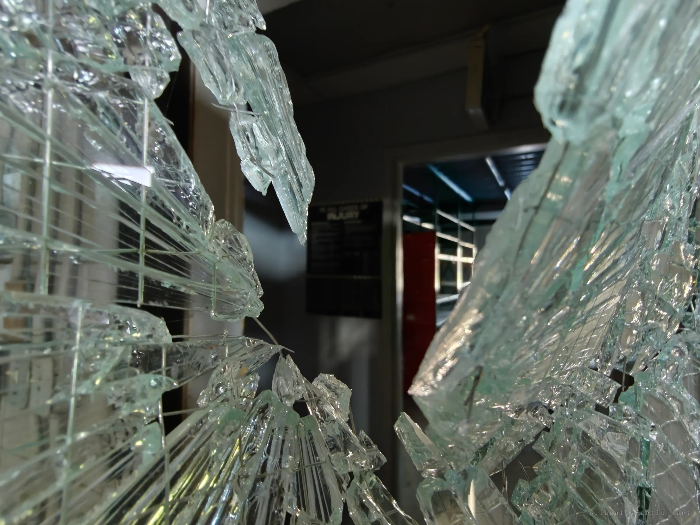 Looking through a shattered pane of safety glass, long splintered shards frame a dim doorway with a black notice board and a red shelf beyond.