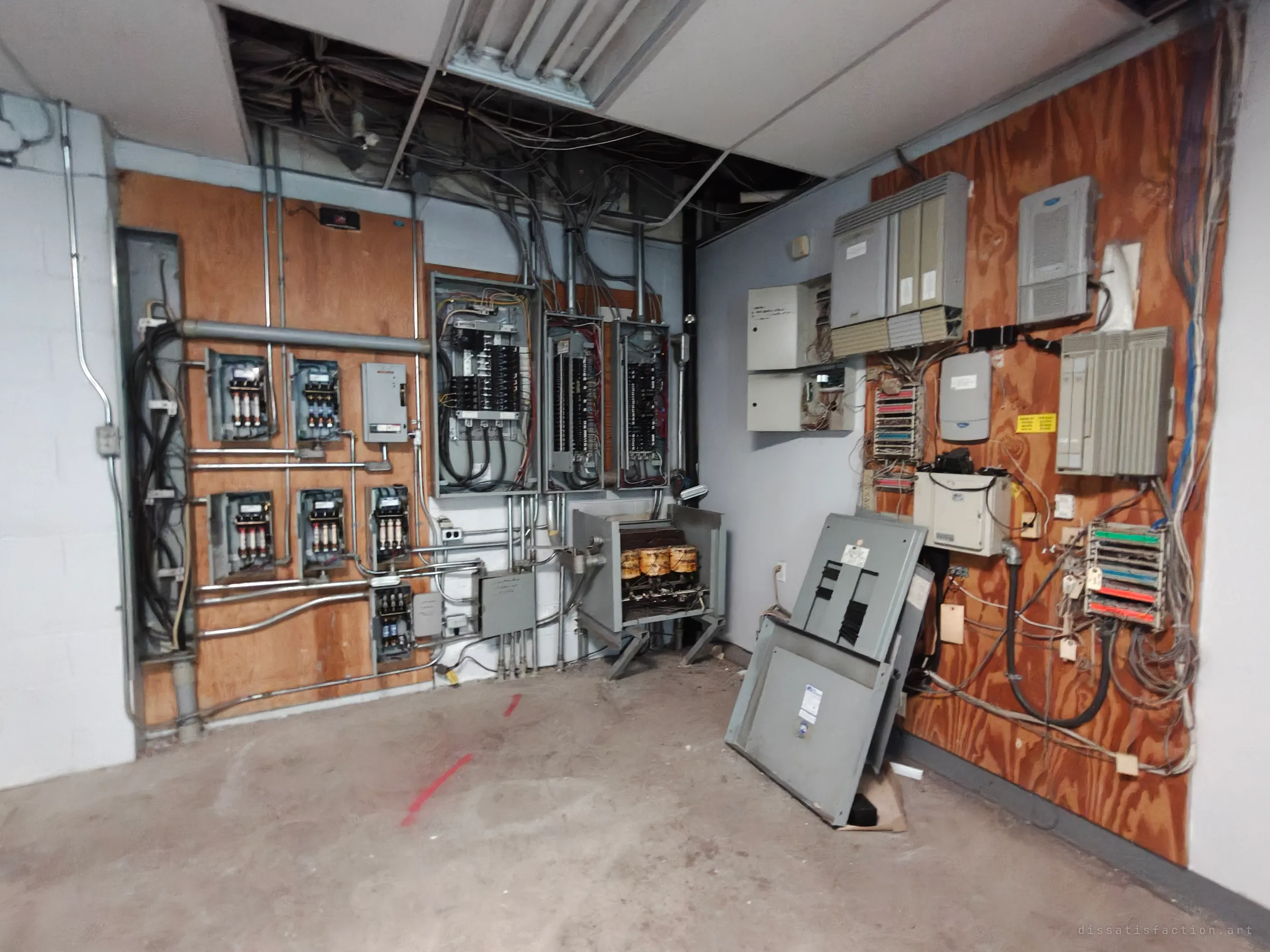 A bare electrical room with fused disconnect switches and breaker panels on one plywood wall and an old tangle of telephone boxes, cables and punch-down blocks on the other, with ceiling tiles pulled away above and a stripped panel cover leaning on the floor.