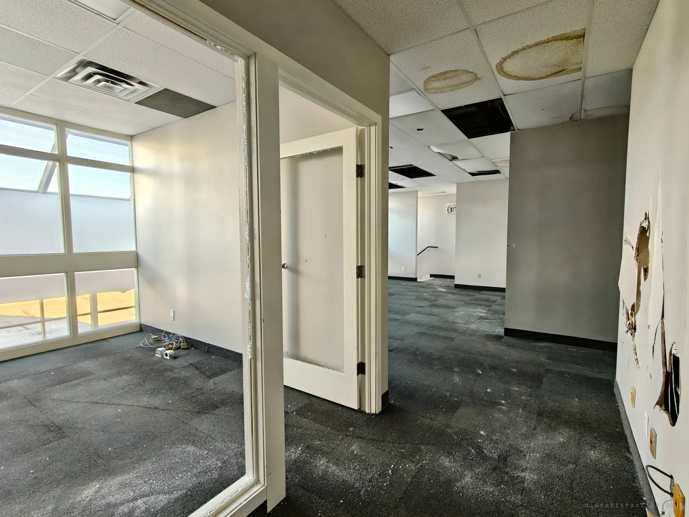 A view through an open doorway into a small glass-walled office with loose cables on the floor. Beyond it, a larger room has stained ceiling tiles and peeling, torn walls.