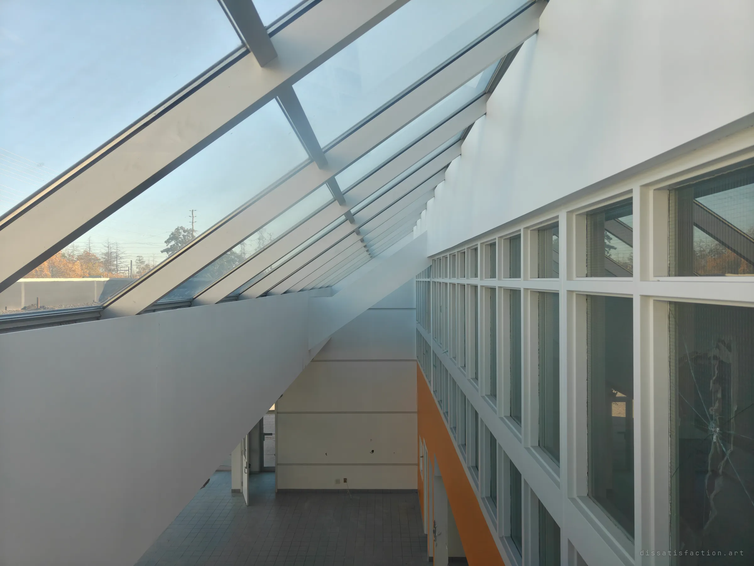 Looking along a sloped glass skylight and a tall white wall of windows down into an empty tiled atrium with an orange wall. The light is low and clean.