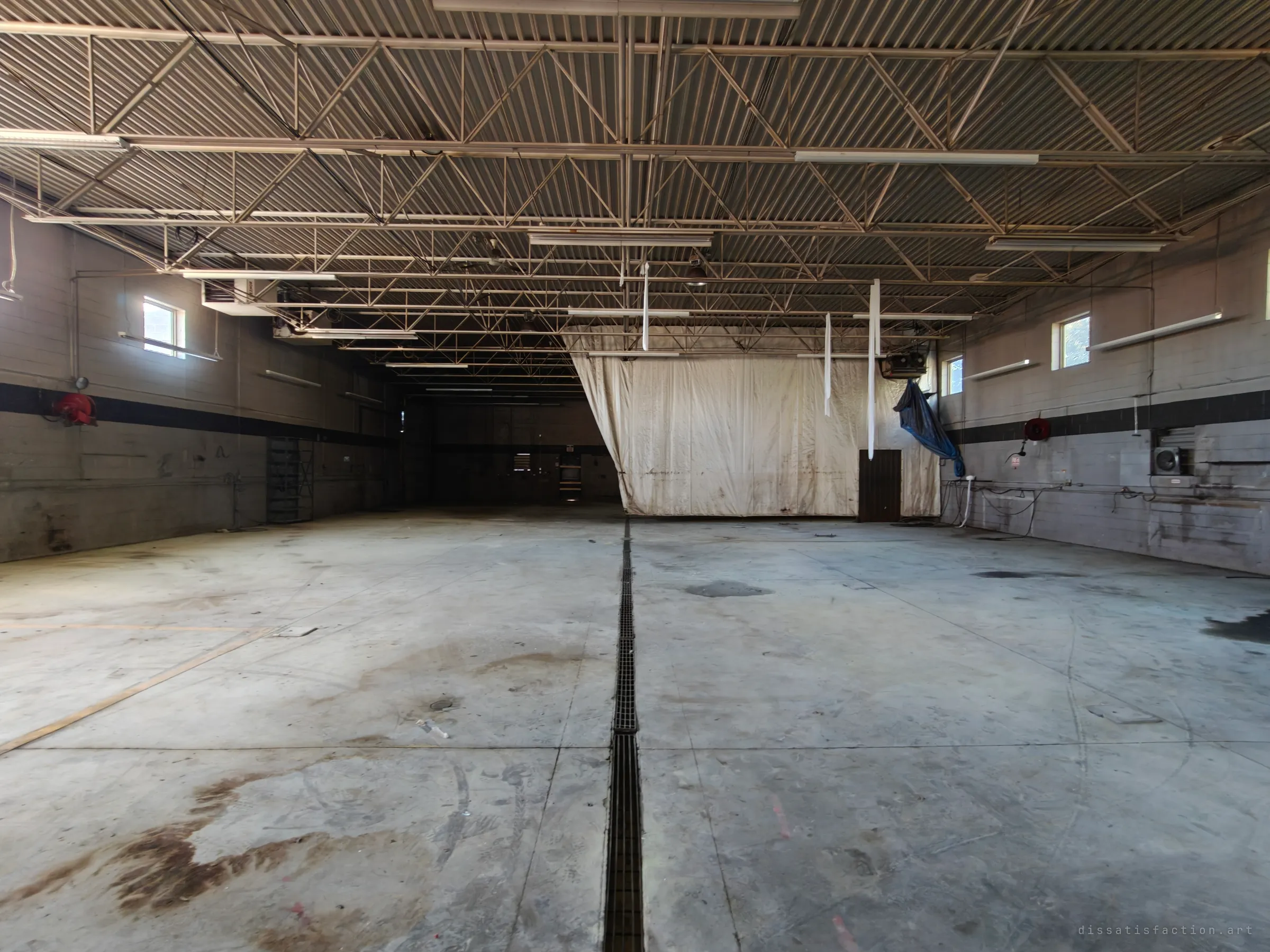 A wide, empty garage bay with a long drain grate running down the middle of a stained concrete floor, steel roof trusses overhead and a dirty hanging curtain at the back.
