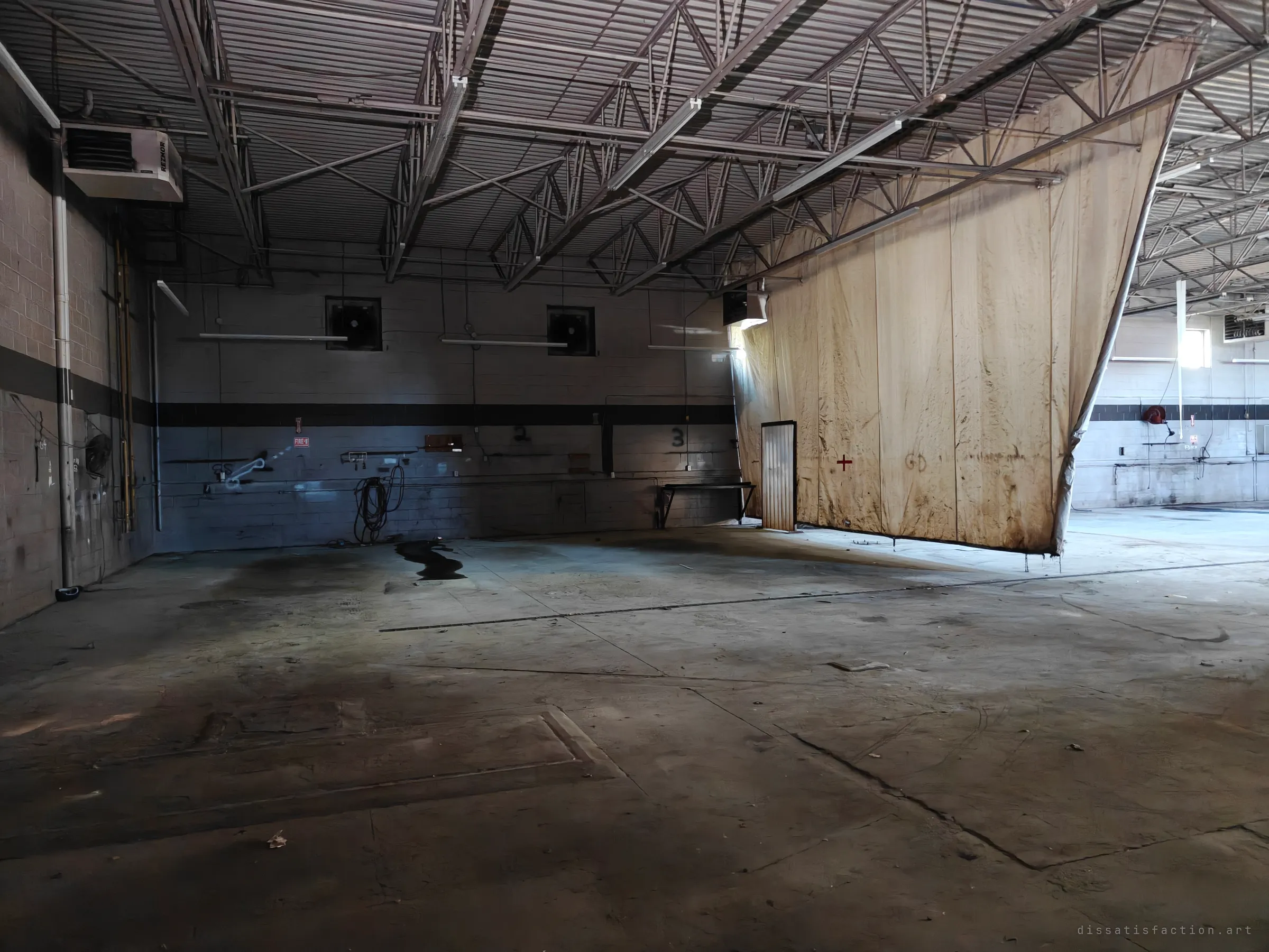 A wide, empty workshop bay under steel trusses, with a huge stained curtain hanging from the ceiling, oil marks on the concrete floor and a heater on the left wall.
