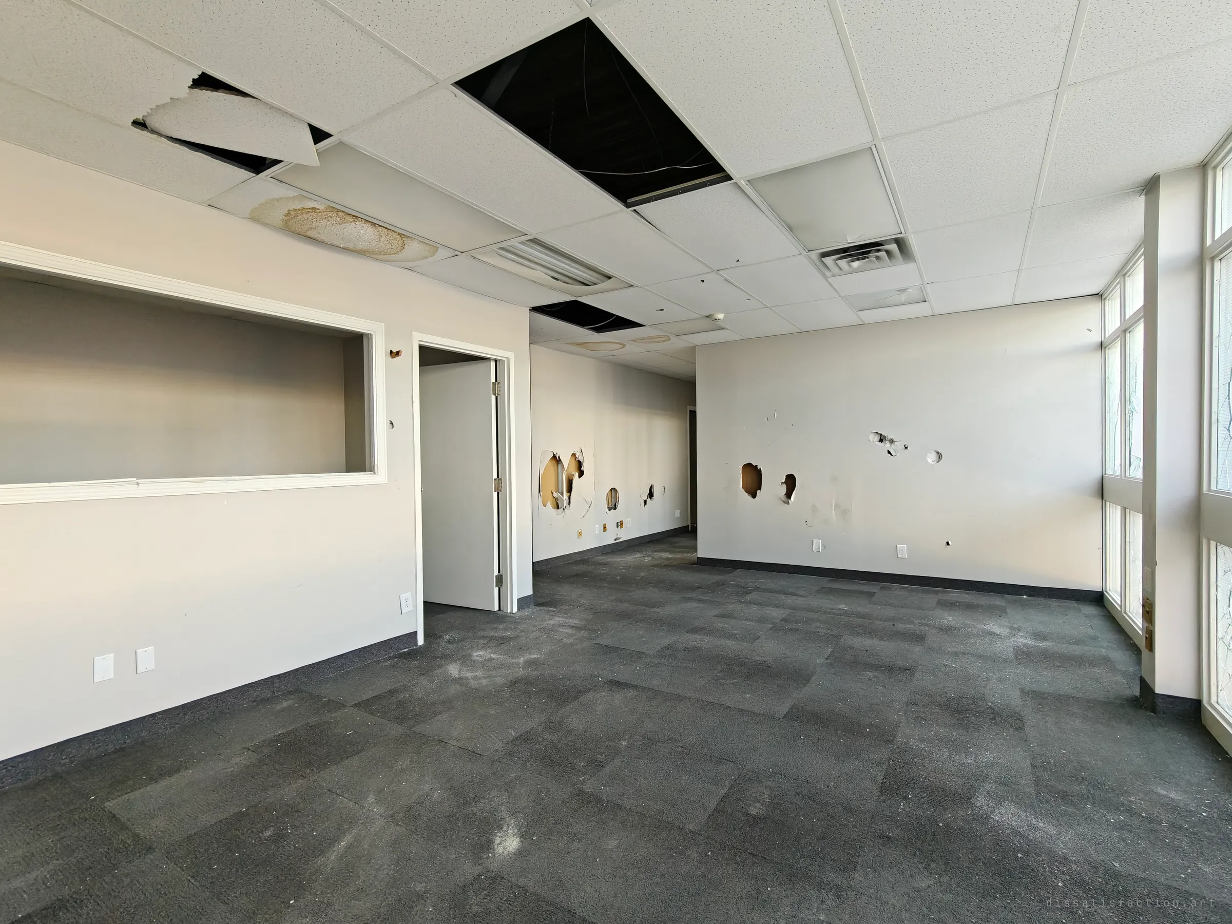 An empty office with water-stained and fallen ceiling tiles. Holes are punched through the walls, an open interior window frame sits on the left, and dusty dark carpet covers the floor.