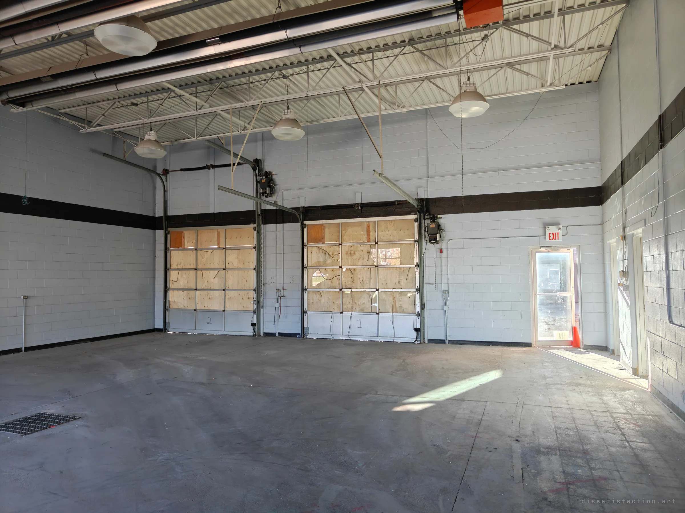 A bare, empty garage bay with two plywood-boarded roll-up doors, white block walls with a black stripe, and sunlight falling across a stained concrete floor.