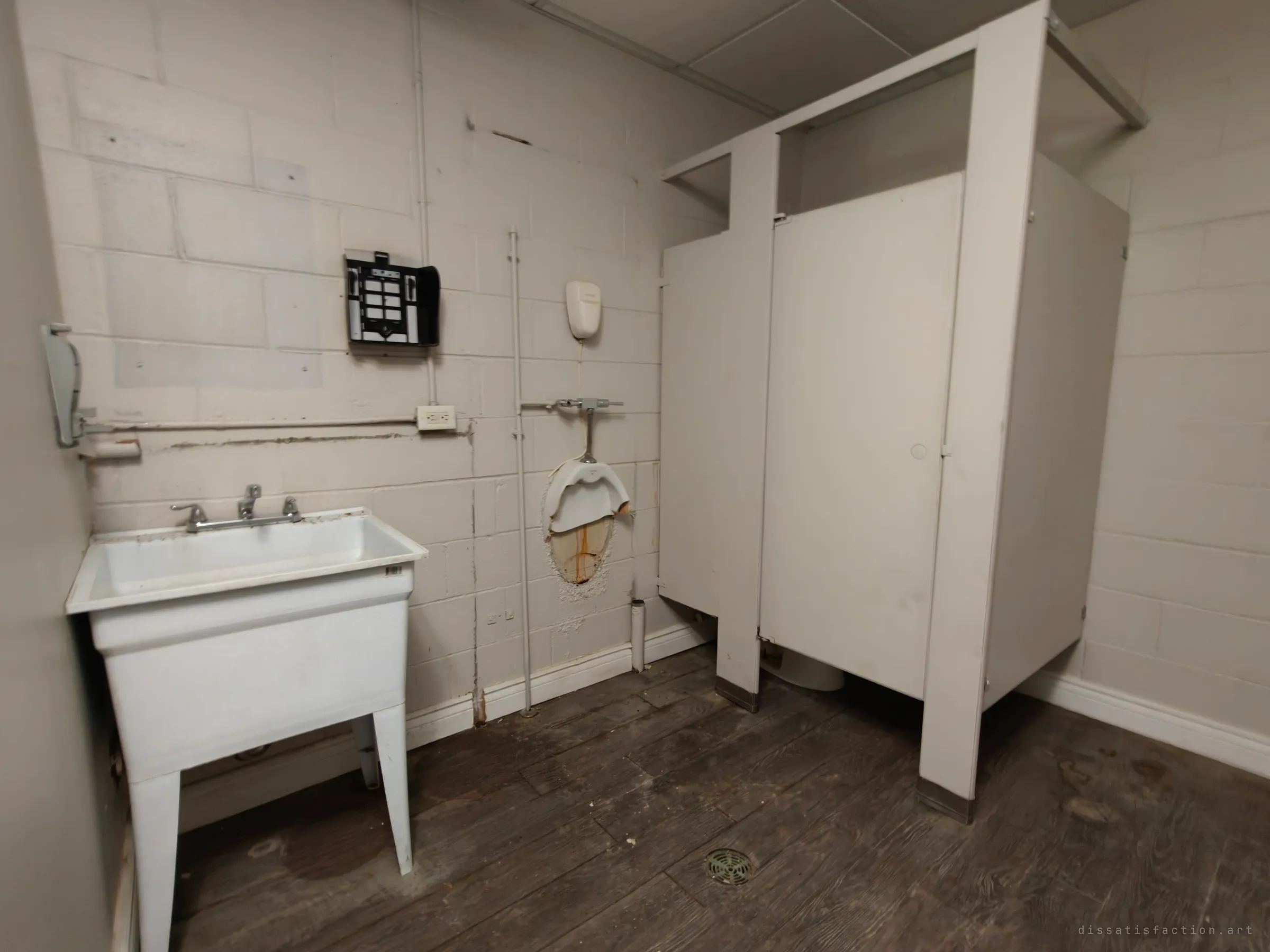 A bare washroom with a white utility sink, a cracked wall-mounted urinal with a rust stain, a standing stall partition and a dark worn plank-style floor.