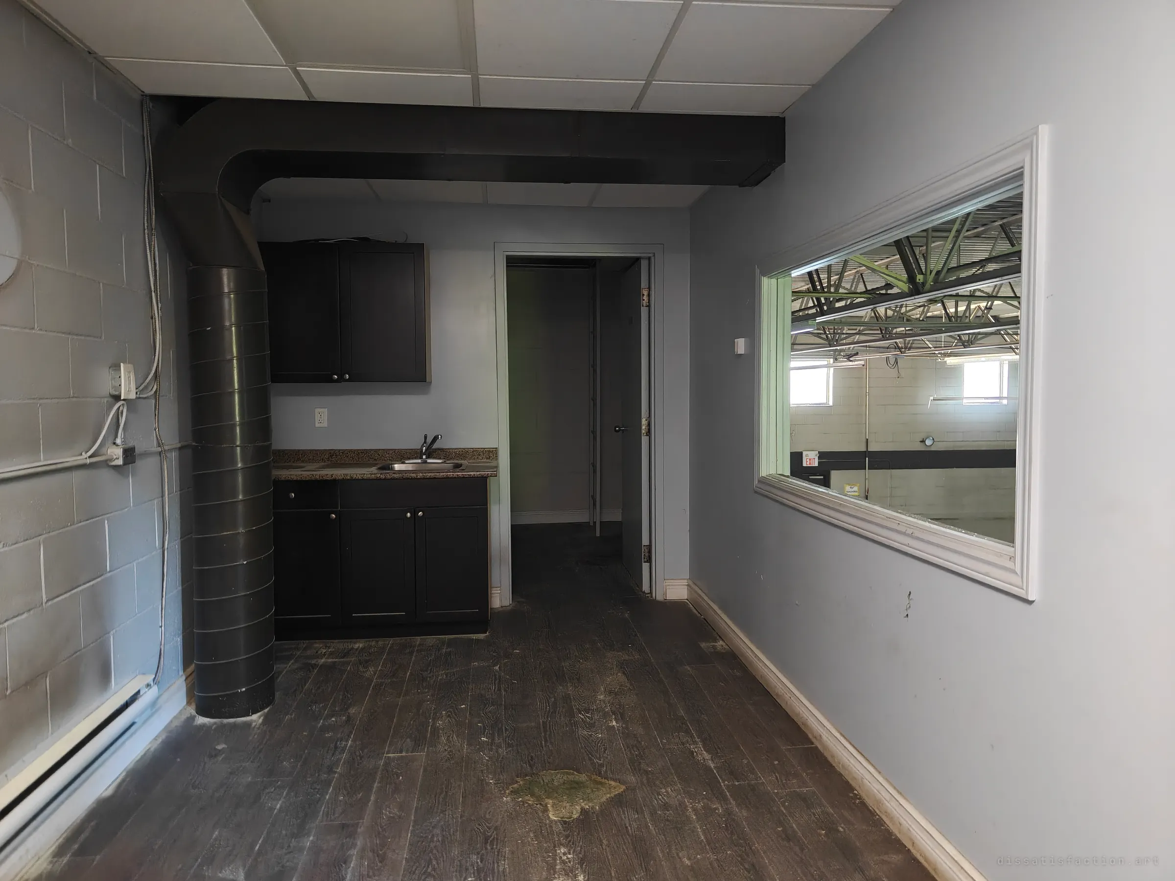 A gutted office kitchenette with dark cabinets, a sink, a large black duct and a stained wood-look floor, and a framed interior window on the right looking out onto a warehouse with green steel trusses.