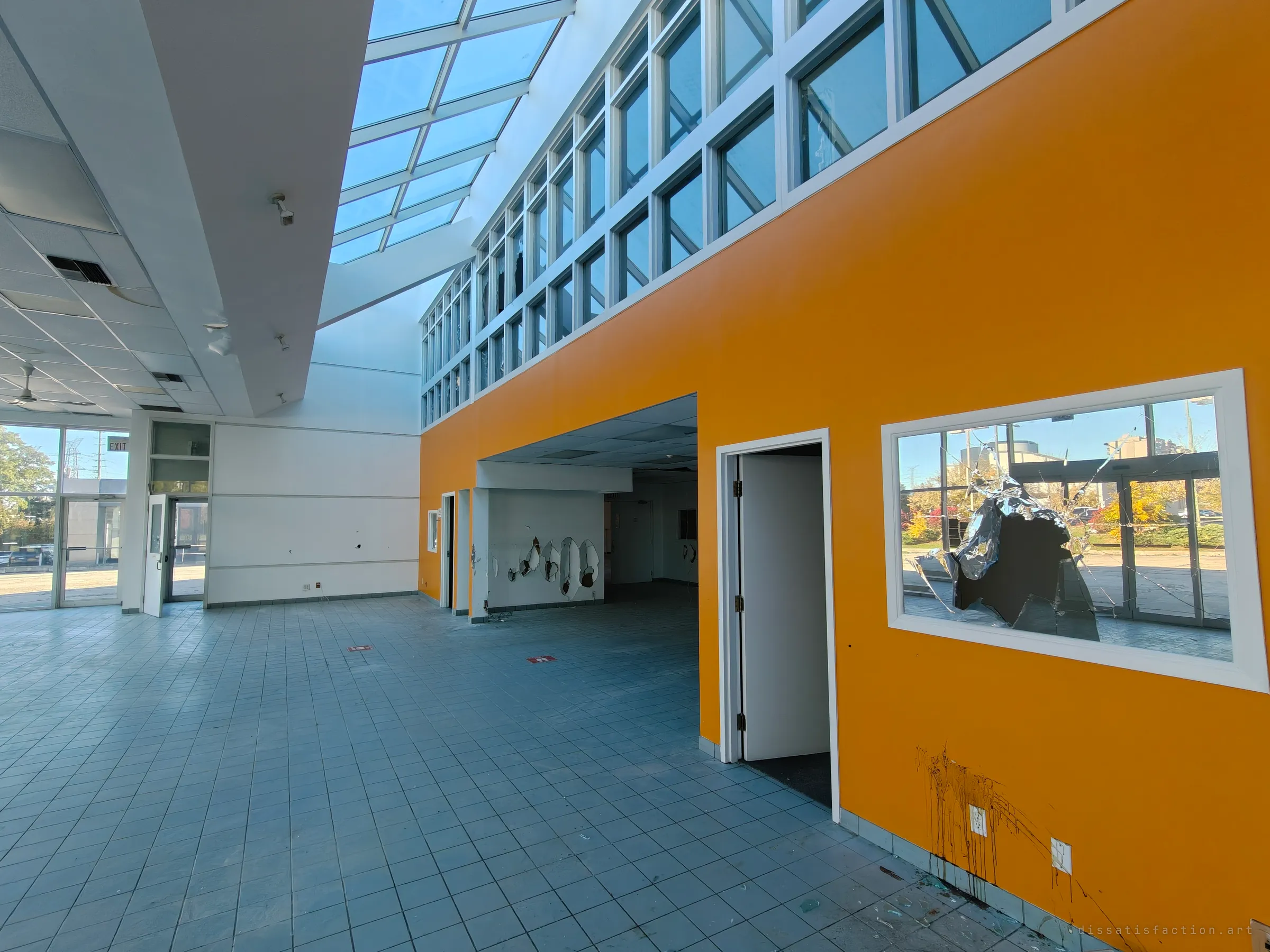 A tall tiled showroom with a glazed skylight and a bright orange wall. A smashed interior window shows a jagged shard of mirrored foil, and brown paint drips down the wall.