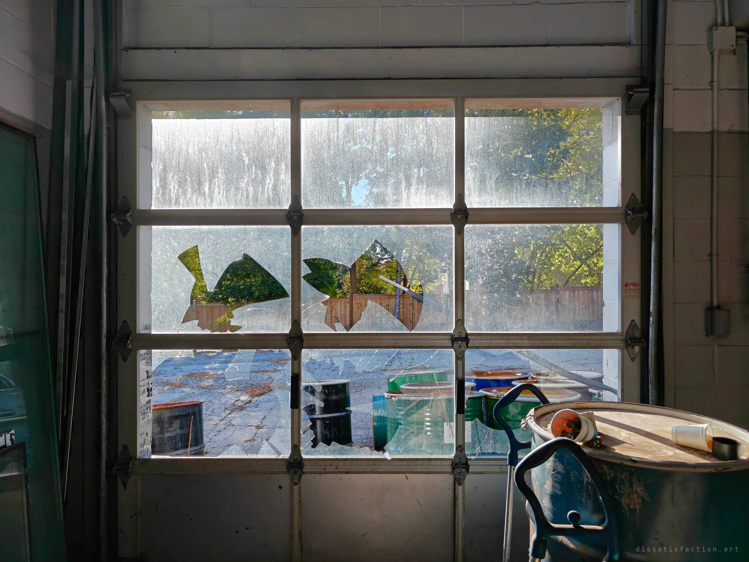 A garage door window with its panes smashed, the shattered holes framing trees, a wooden fence and a yard of old drums, backlit by hard daylight, with a drum in the foreground.