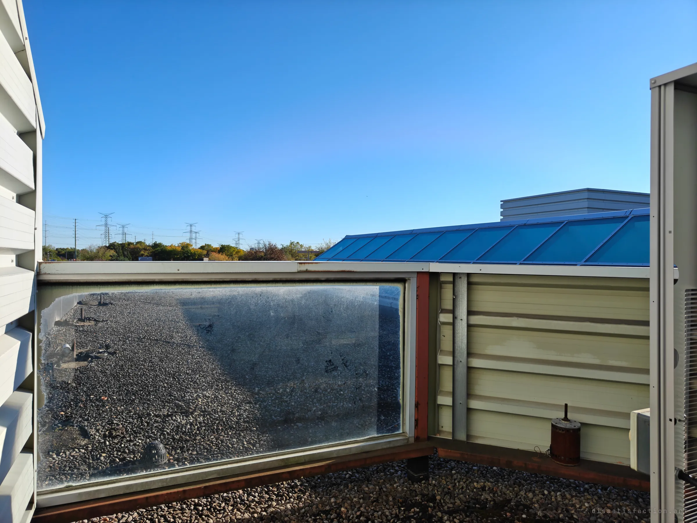 A rooftop seen between a wall of louvres and an air-conditioning unit. A grimy glass panel looks out over gravel ballast and a roof drain, with a blue skylight and pylons in the distance.