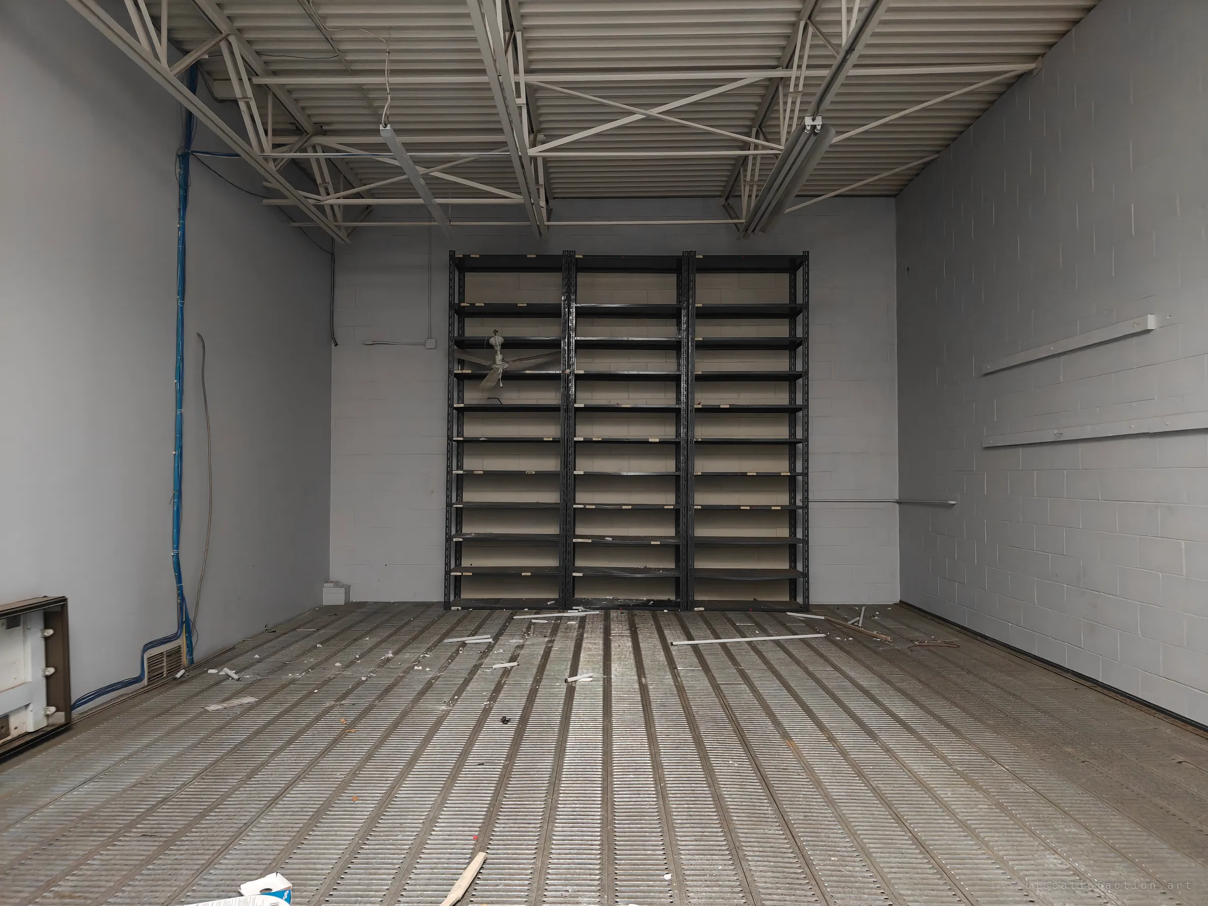 A tall empty room with a steel-truss ceiling and a metal grating floor scattered with debris, with three bays of black shelving against the far wall and a blue cable running down the left wall.