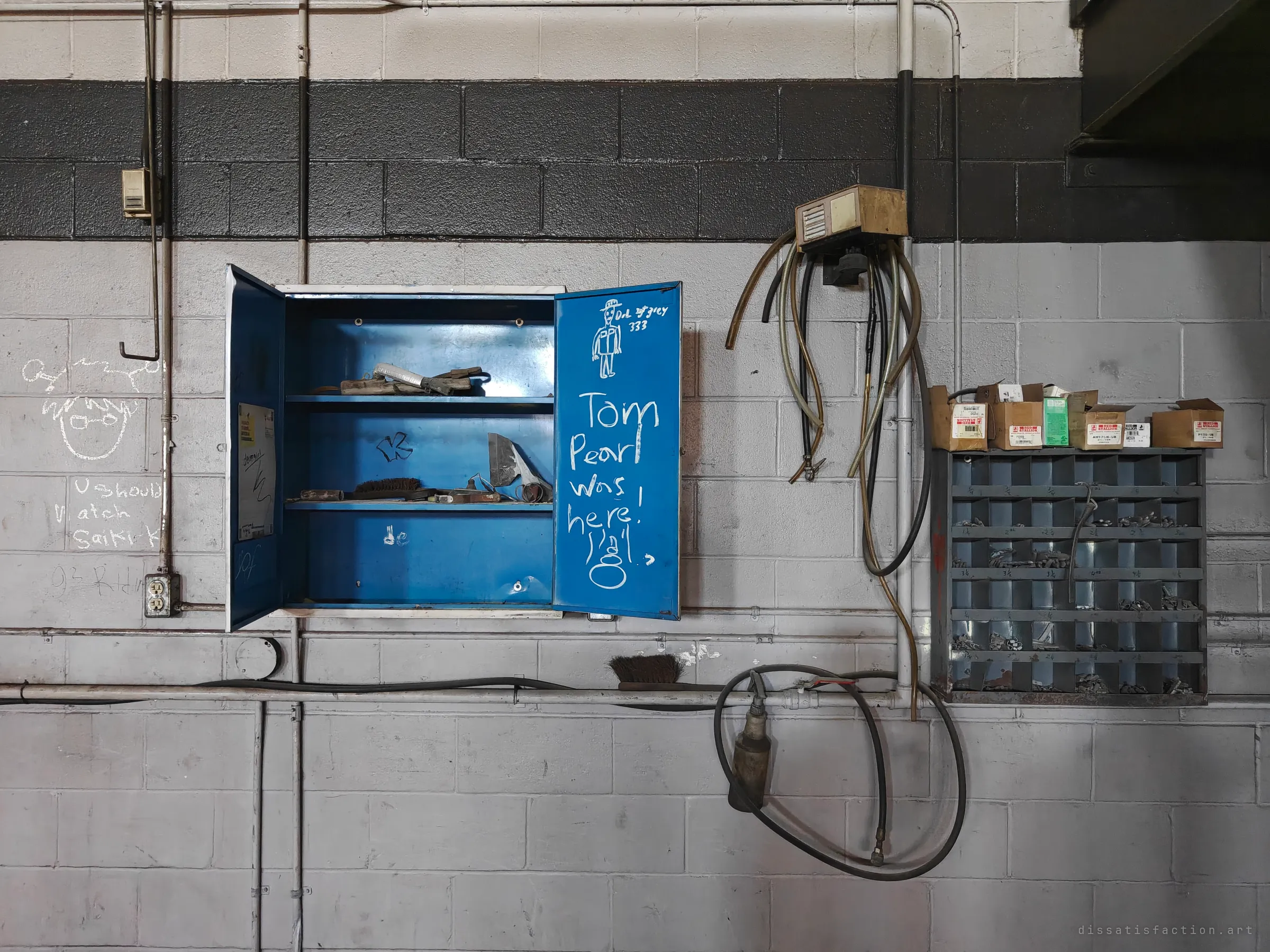 A blue wall cabinet hangs open on a grey block wall, with chalk graffiti on its door and a small drawing of a figure above it. Hoses and a bin of loose hardware hang beside it.