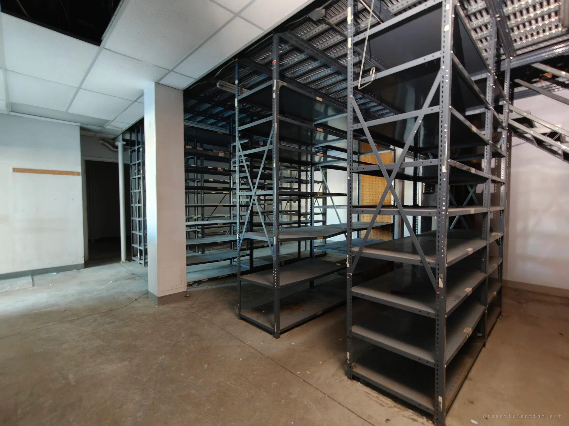 Rows of empty grey steel shelving stand under a perforated metal mezzanine and stair in a dim storage room, with bare floor and a dark doorway at the left.