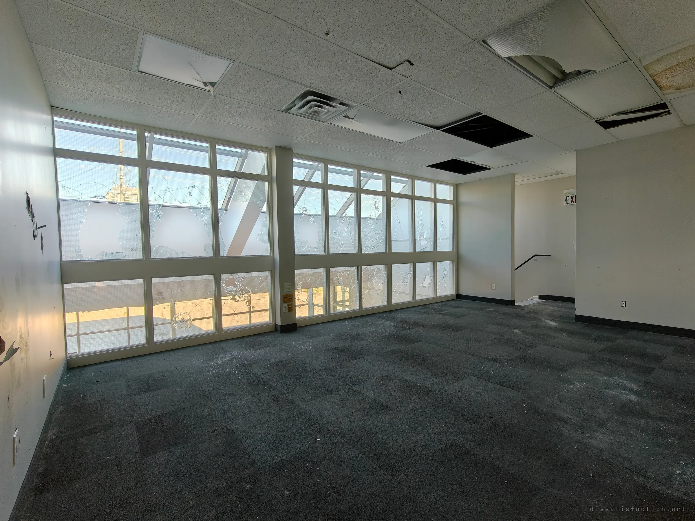 A bare office room with a dark, dusty carpet and a wall of smashed windows. Cracked glass lets in daylight, and ceiling tiles hang open above.