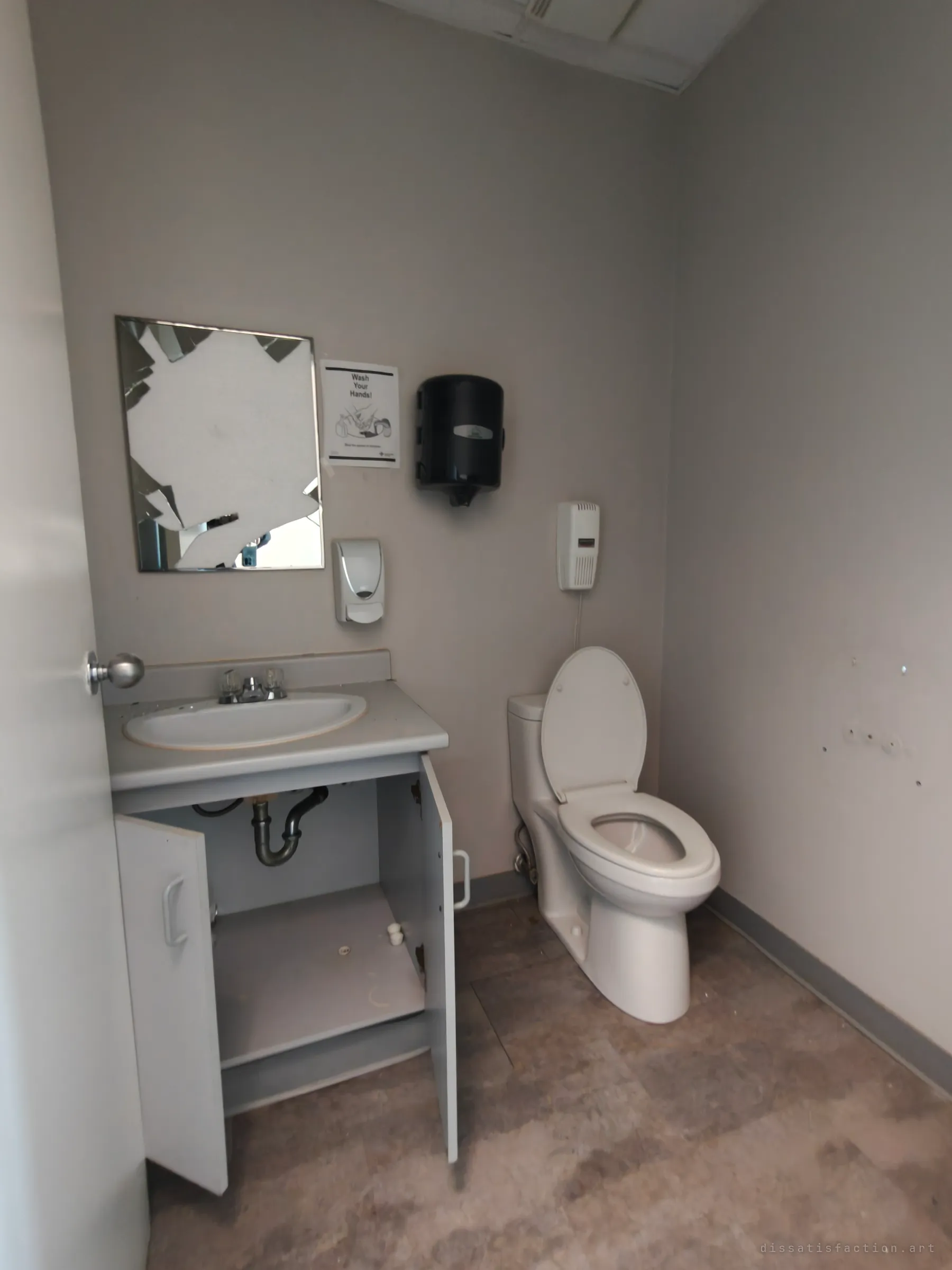 A small bathroom stripped back to the basics. A smashed mirror hangs over a sink with its cabinet doors open, a hand-washing poster is pinned beside it, and a toilet sits on a grimy floor.