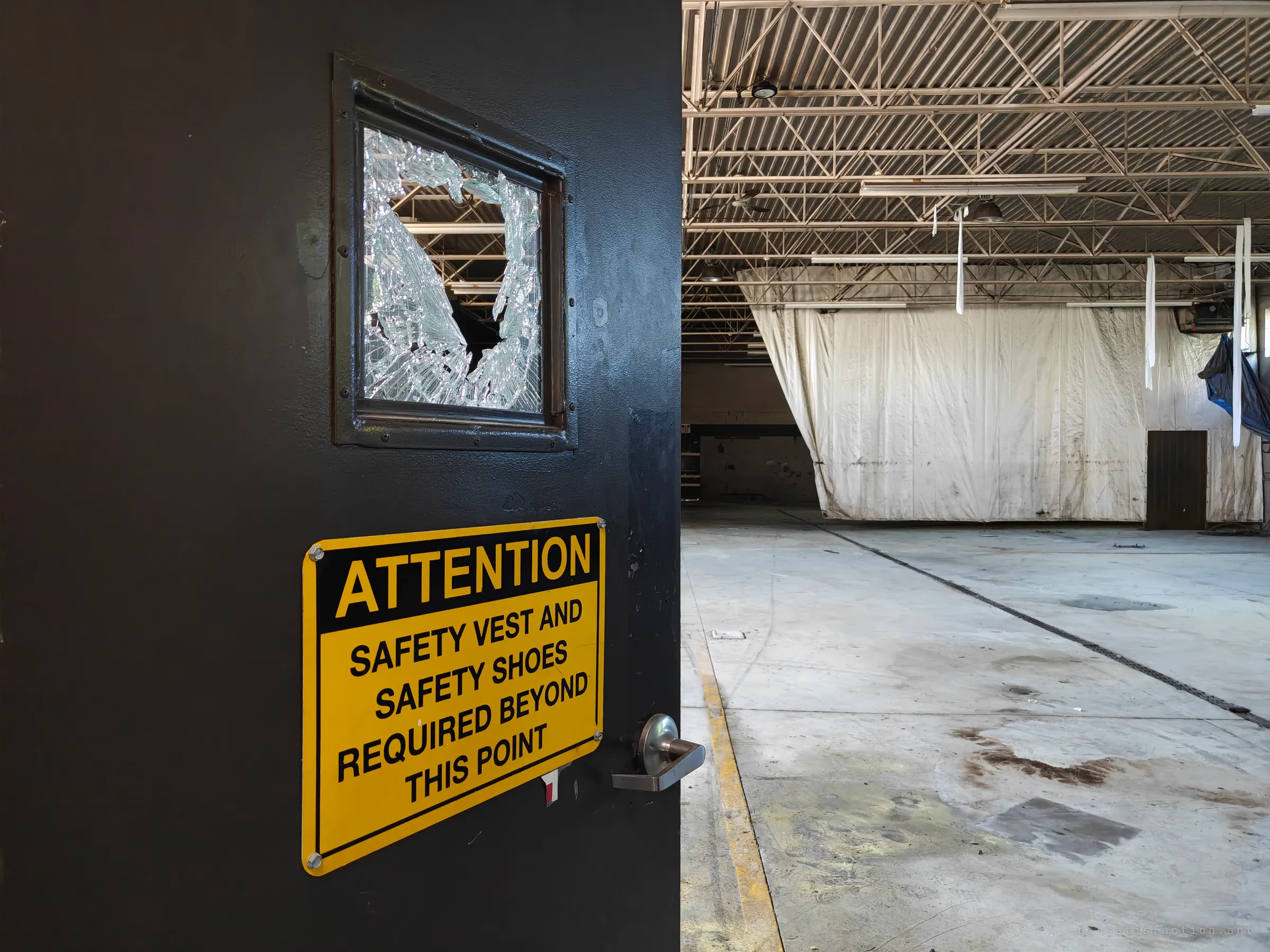 A black door with a smashed window and a yellow safety-vest-and-shoes warning sign opens onto a large empty hall, with a sheeted-off partition and bare roof trusses beyond.