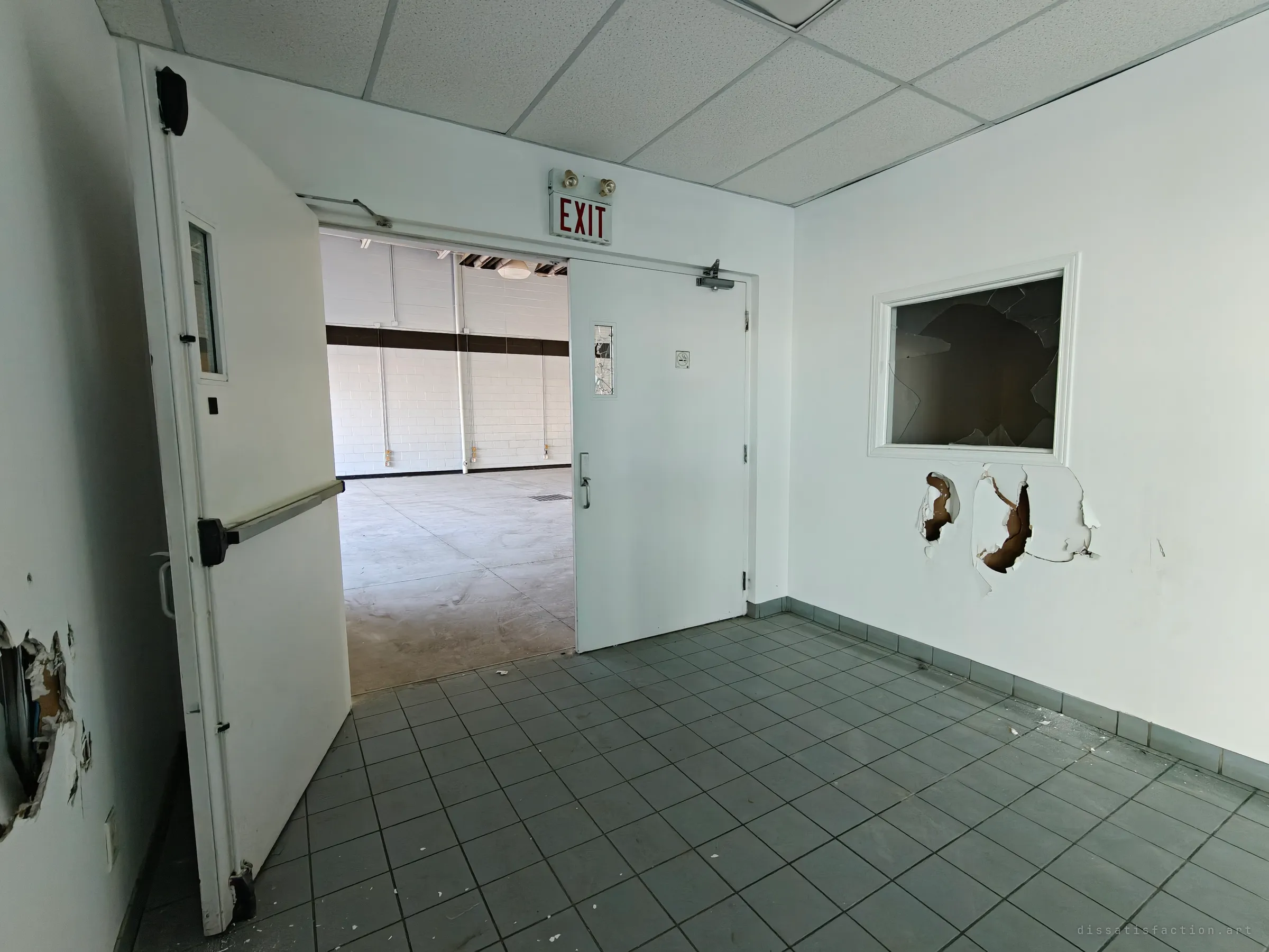 A tiled entry with a propped double door opening onto an empty, stripped floor beyond, and a smashed interior window and torn drywall on the right wall under an exit sign.