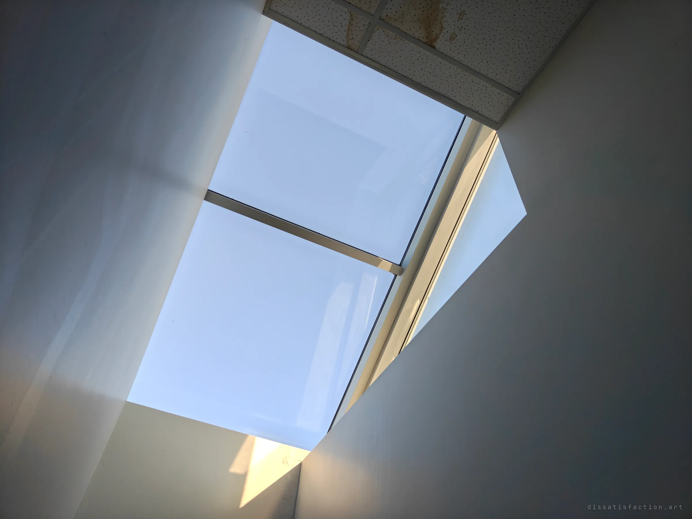 Looking up a bare white shaft at a skylight, its glass washed pale blue, with a water-stained ceiling tile at the top edge.