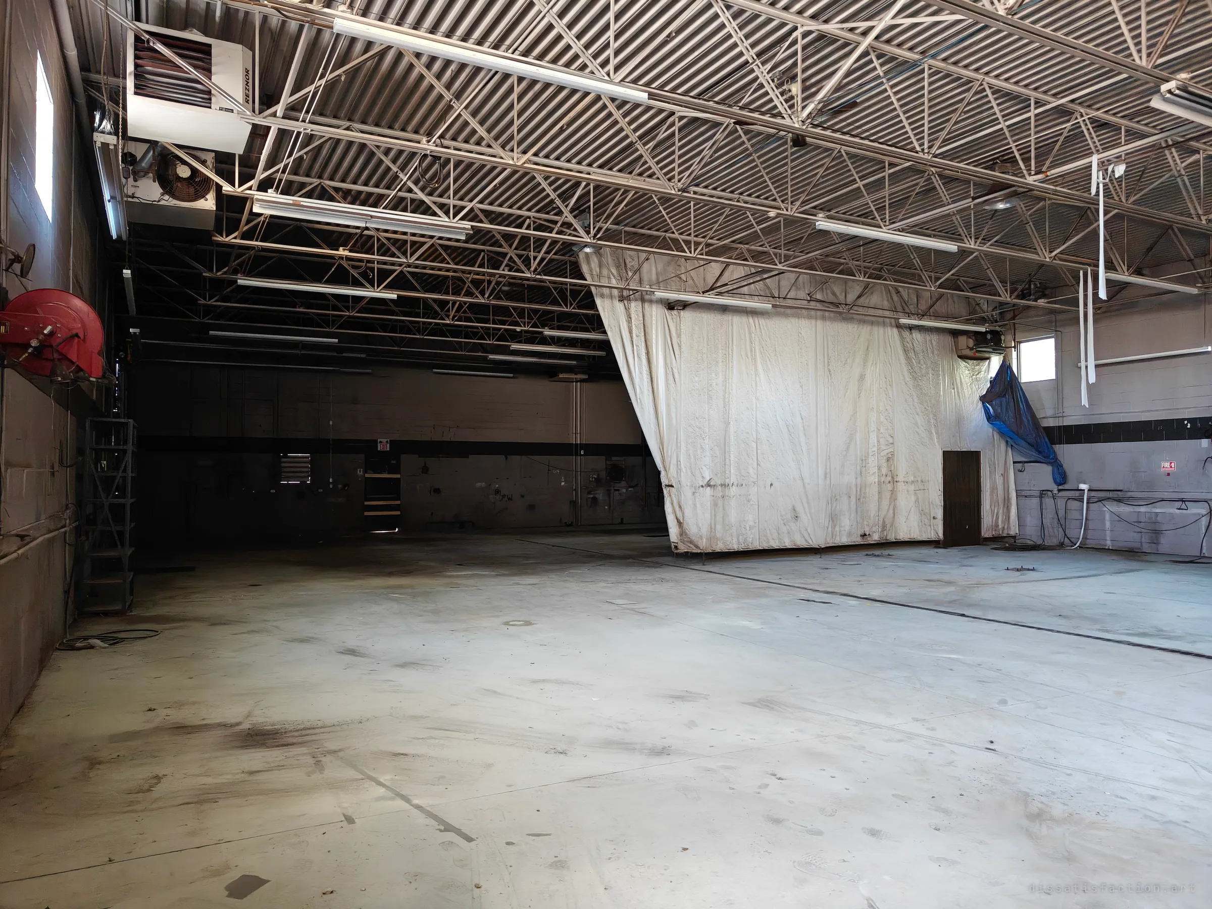 Corner of an empty steel-trussed garage bay, with a red hose reel on the left, a ceiling heater and a stained white curtain hanging across the far side.