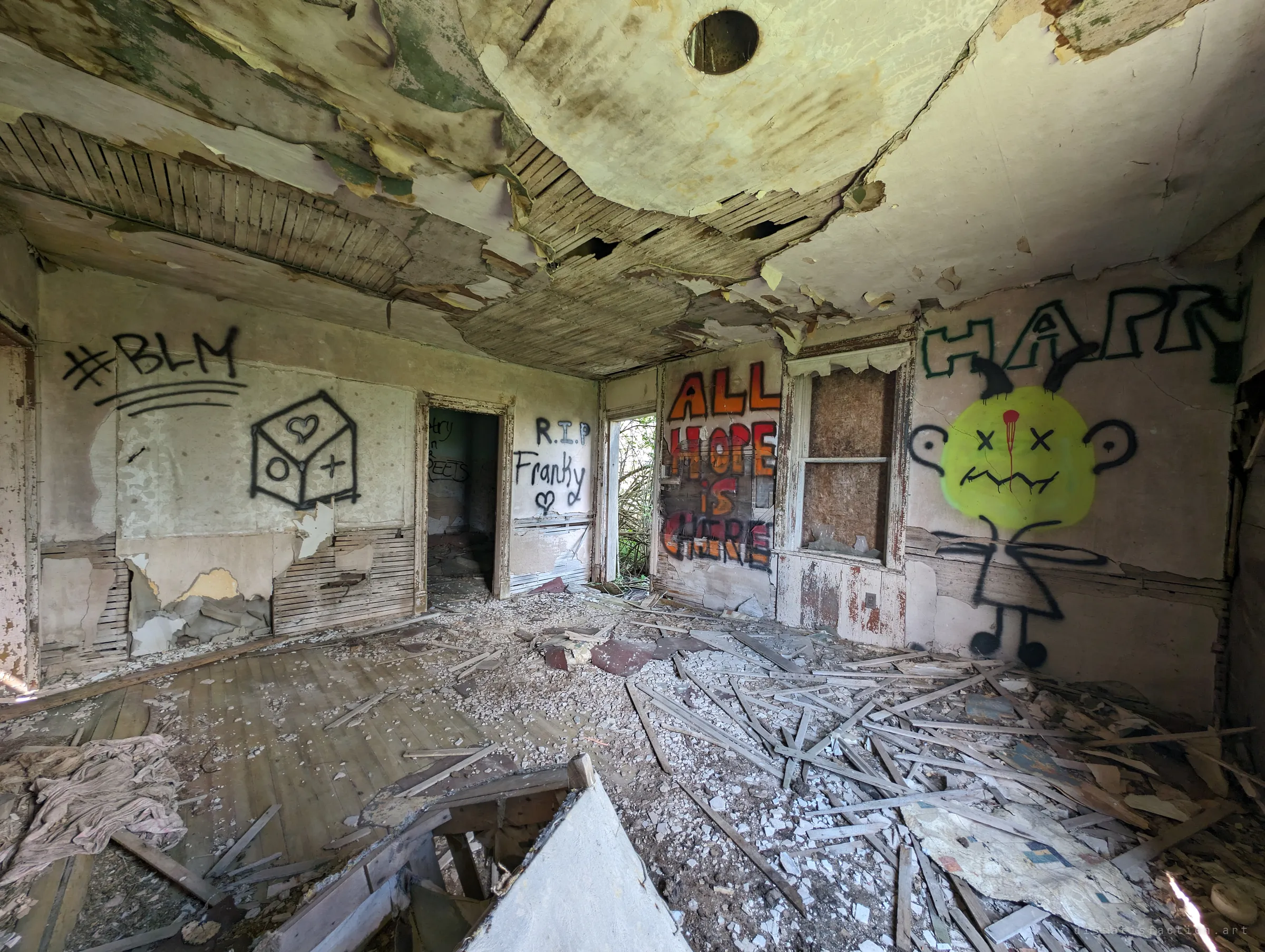 A wide view of the main room: the ceiling plaster hangs in sheets over exposed lath, the floor is buried in debris, and every wall carries graffiti, including a bright yellow cartoon face and the words "all hope is here". It captures how thoroughly the house has been visited and wrecked.