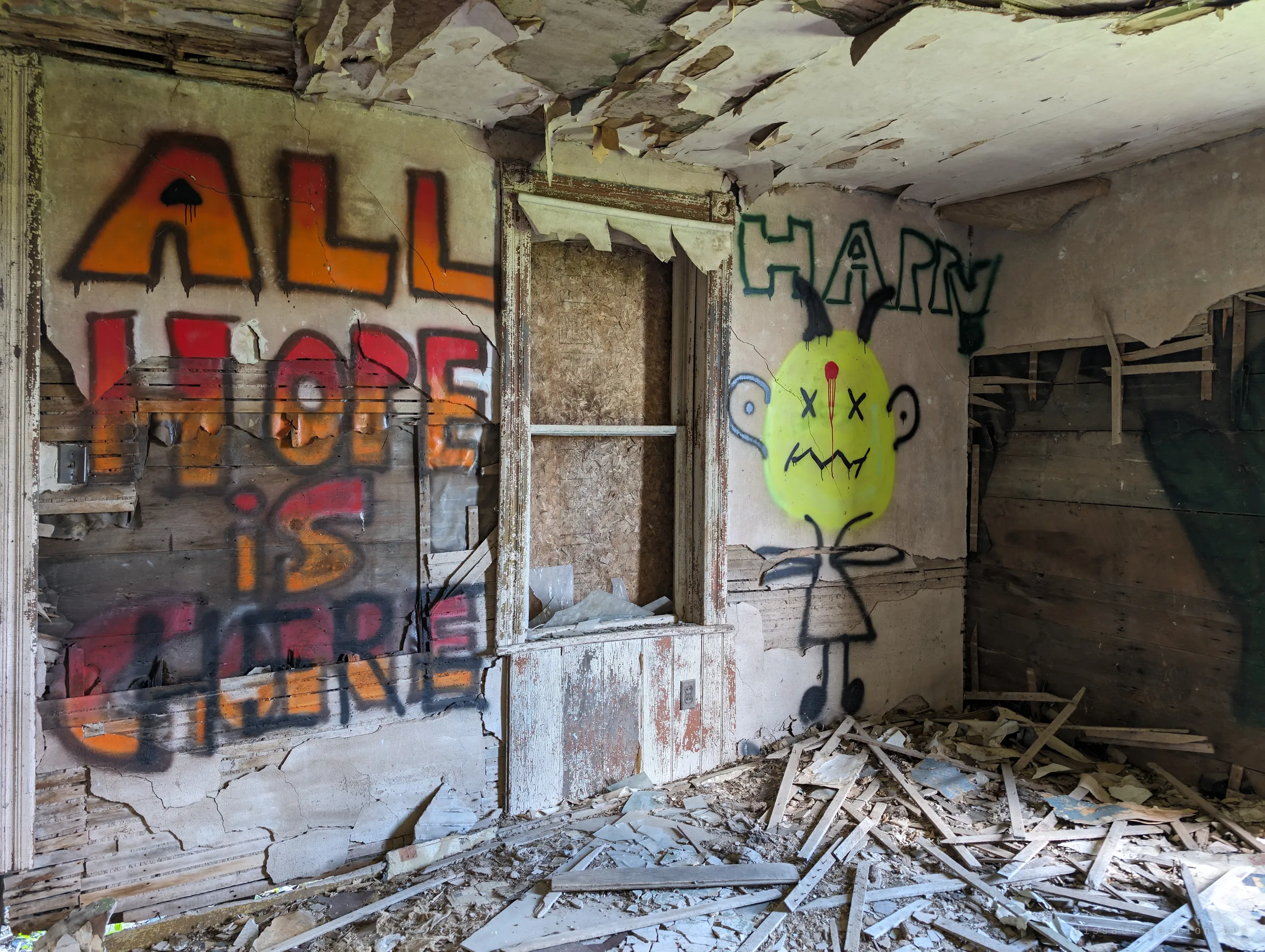 Closer on the graffiti wall: "all hope is here" in dripping orange and red letters beside a boarded built-in cabinet and a yellow cartoon creature with crossed-out eyes. Lath and broken plaster pile up on the floor below.