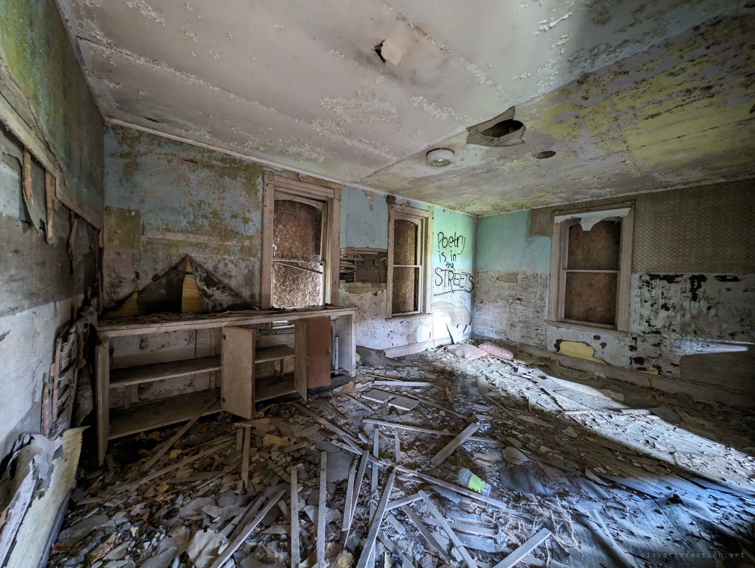A former kitchen with open wooden cupboards, boarded windows and pale blue walls peeling to plaster, the floor covered in fallen lath. Sunlight slants across the debris next to a scrawled line about poetry in the streets.