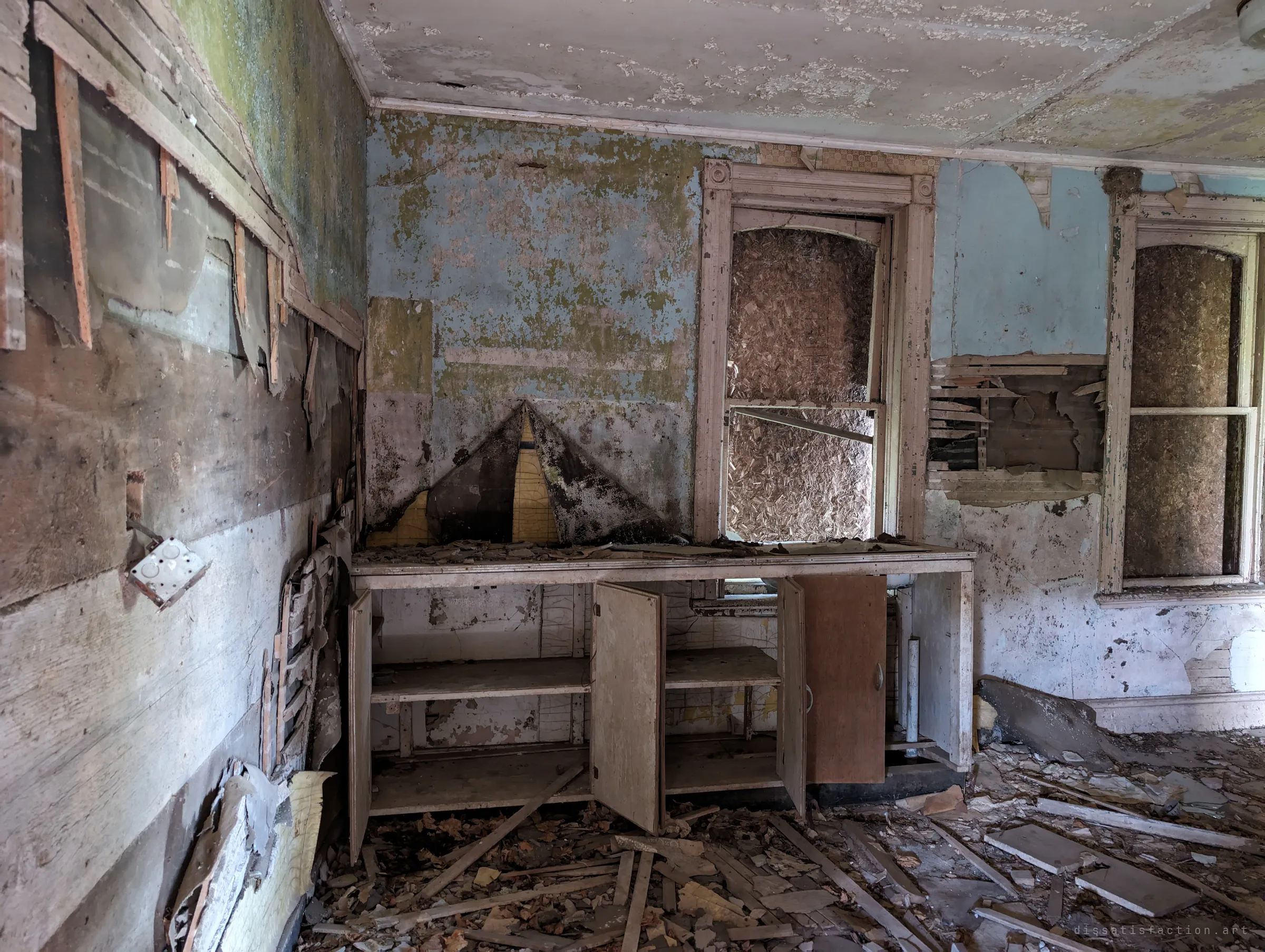 A gutted kitchen corner with open, empty cabinets beneath a boarded-up window, pale blue paint and wallpaper peeling off the walls in sheets. Debris and broken lath cover the floor.