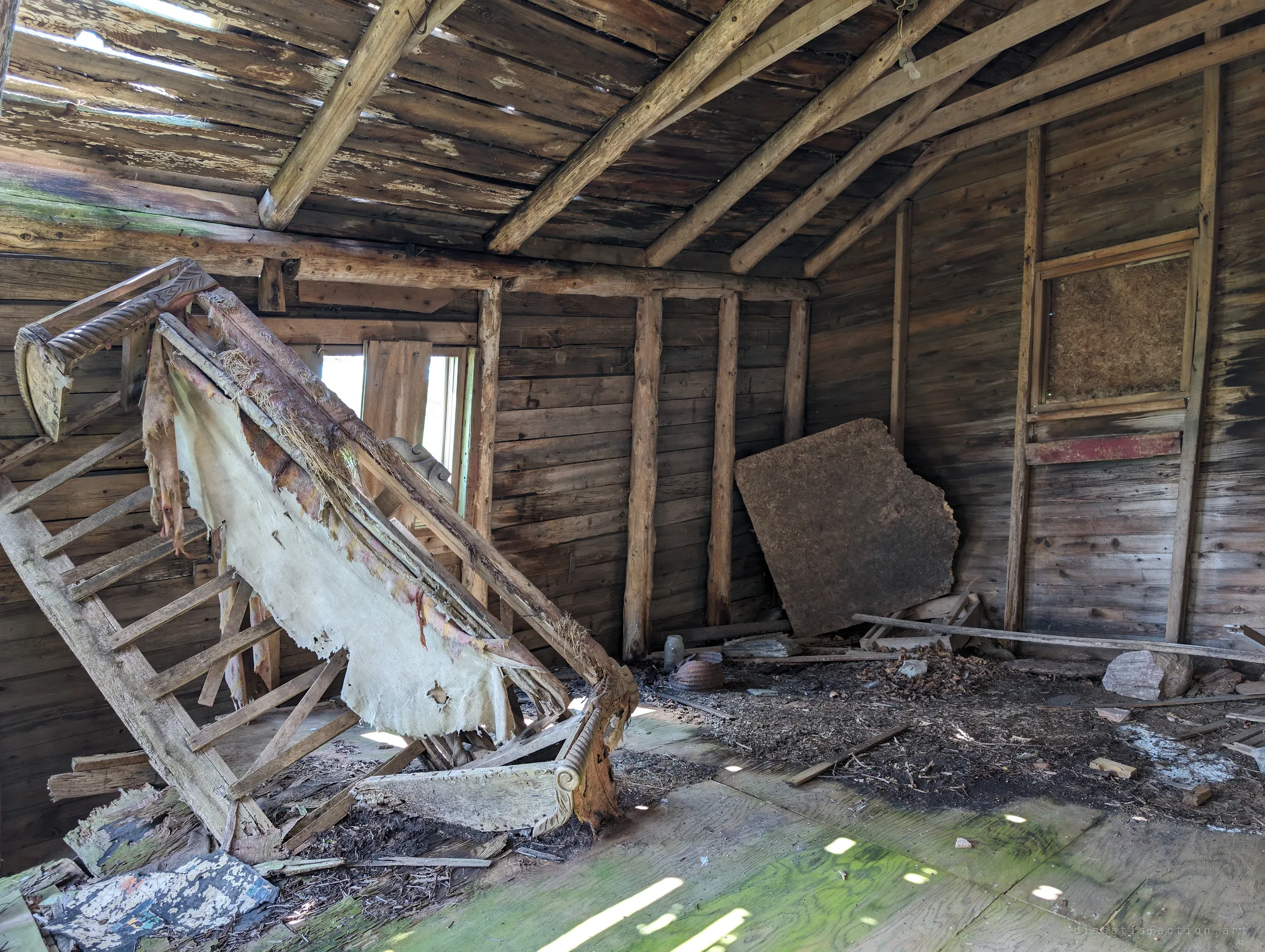 A rough log-framed upper room where a torn, ornately carved sofa frame leans on its end against a ladder. Light slips through gaps in the plank roof onto a mossy floor.