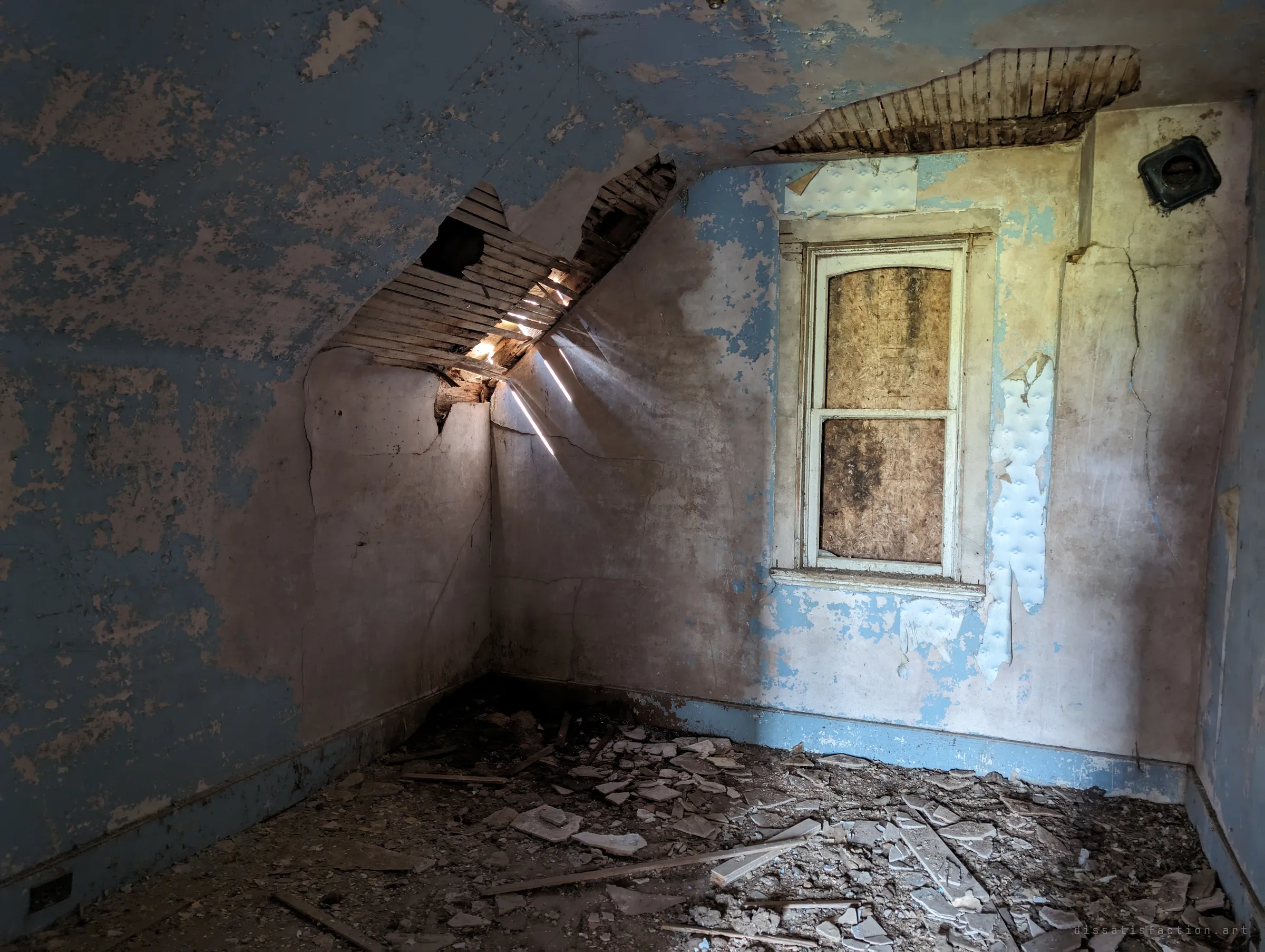 A small blue attic bedroom with a boarded window, where a hole in the sloped ceiling lets thin beams of sunlight cut across the peeling plaster. Broken plaster litters the floor.