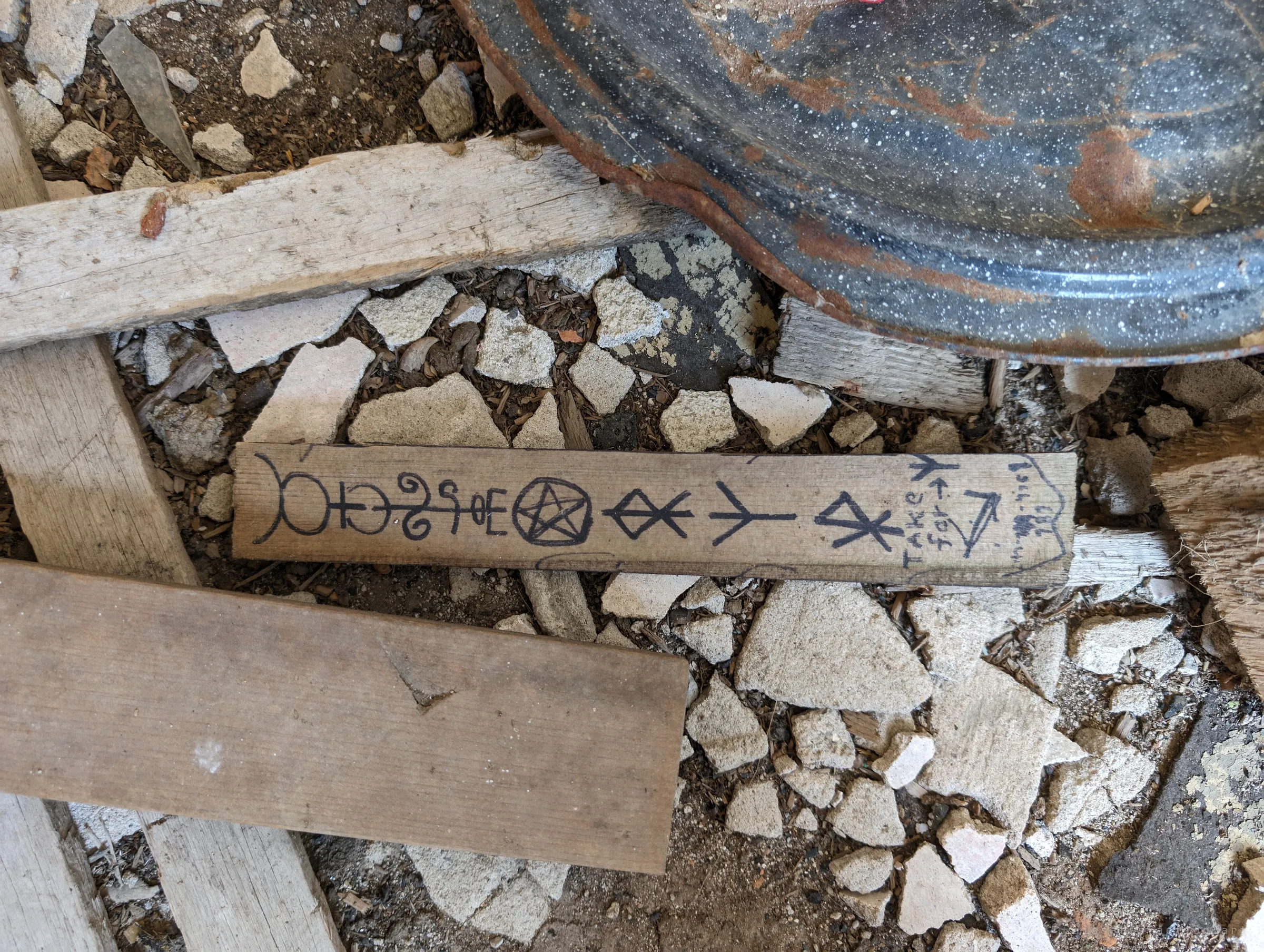A strip of wood lying in plaster rubble, marked in black ink with a row of occult symbols, runes and a pentagram. Left behind by an earlier visitor, it hints at the house's later life as an informal shrine.