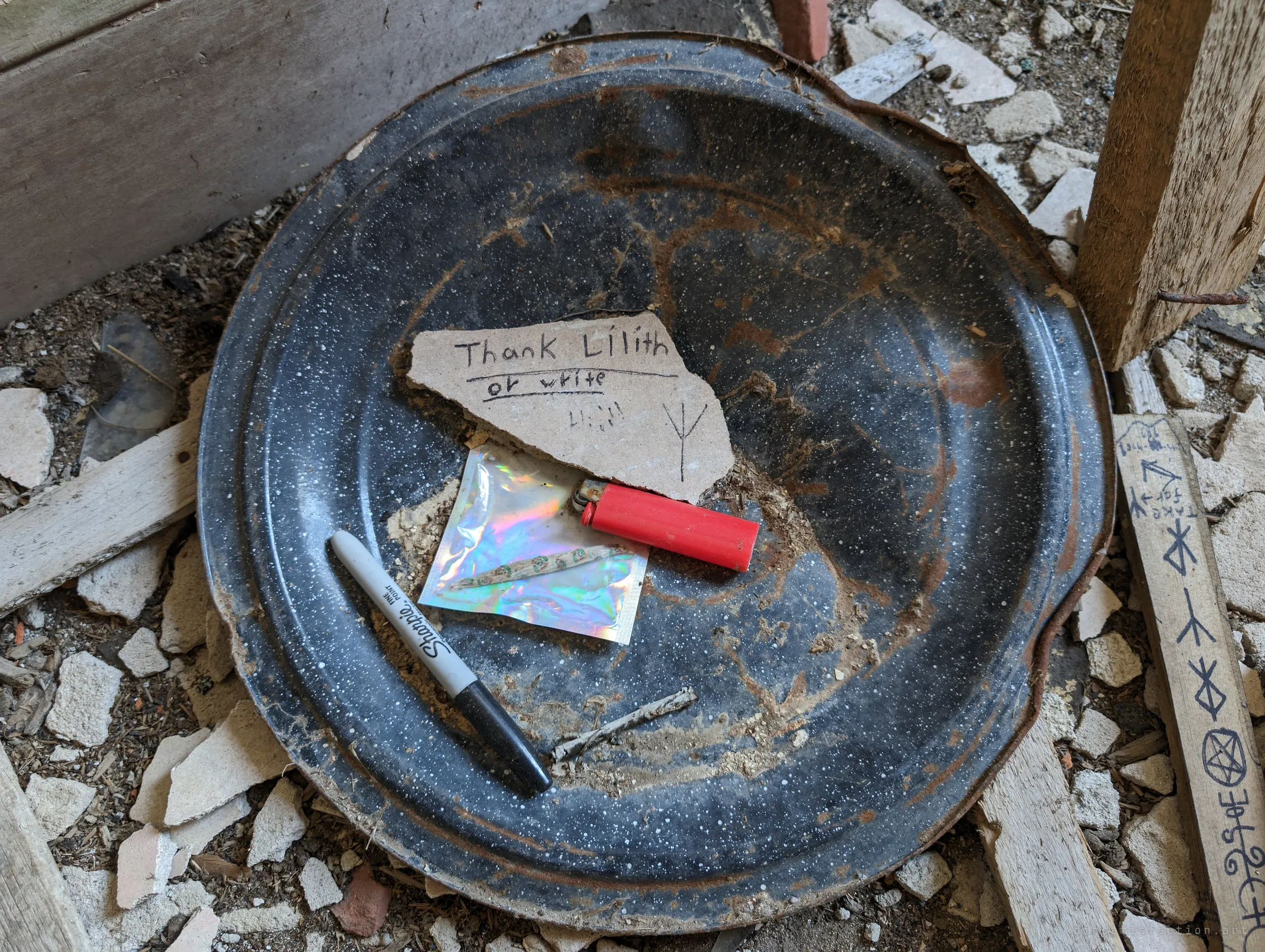 An old enamel basin set up as an offering dish, holding a plaster shard with a handwritten note to a mythological figure, a lighter, a marker and a small foil packet. A rune-marked stick lies beside it.