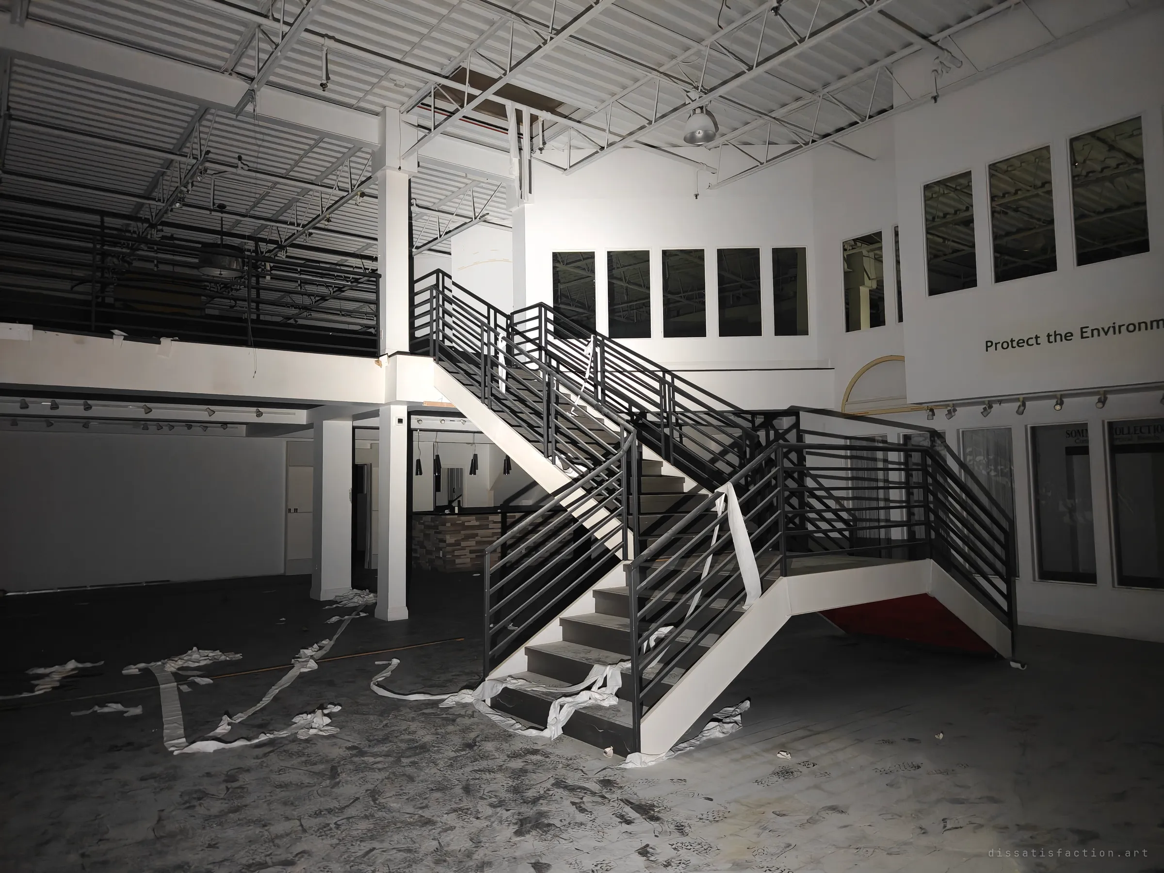 A floating steel staircase rises to a mezzanine in the empty white showroom, toilet paper strung along the railings and trailing across a floor covered in footprints. The design-showroom finish contrasts with the mess left behind.