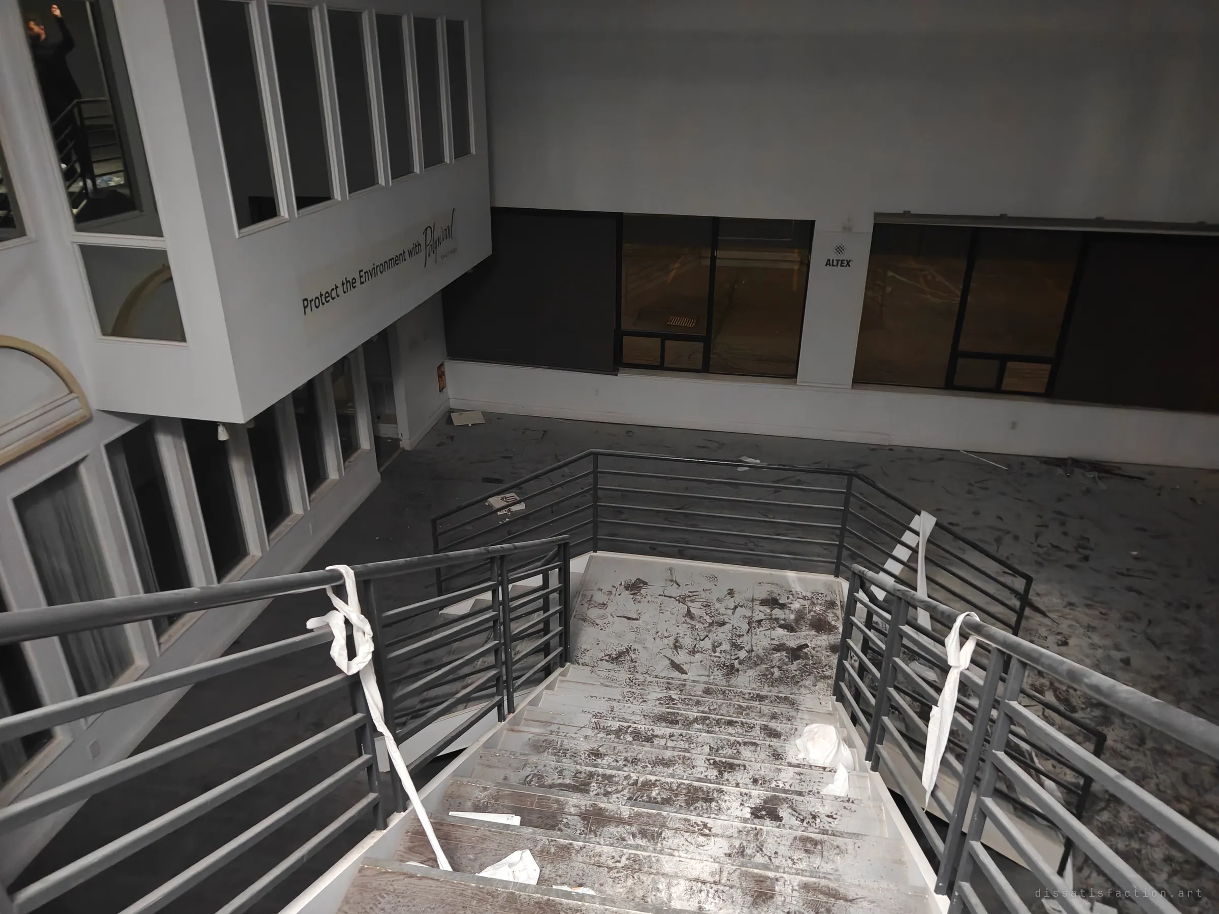 Looking down the showroom stairs into a lower hall, the steps smeared with dirty footprints and toilet paper tied to the rails. Dark windows line the far wall.