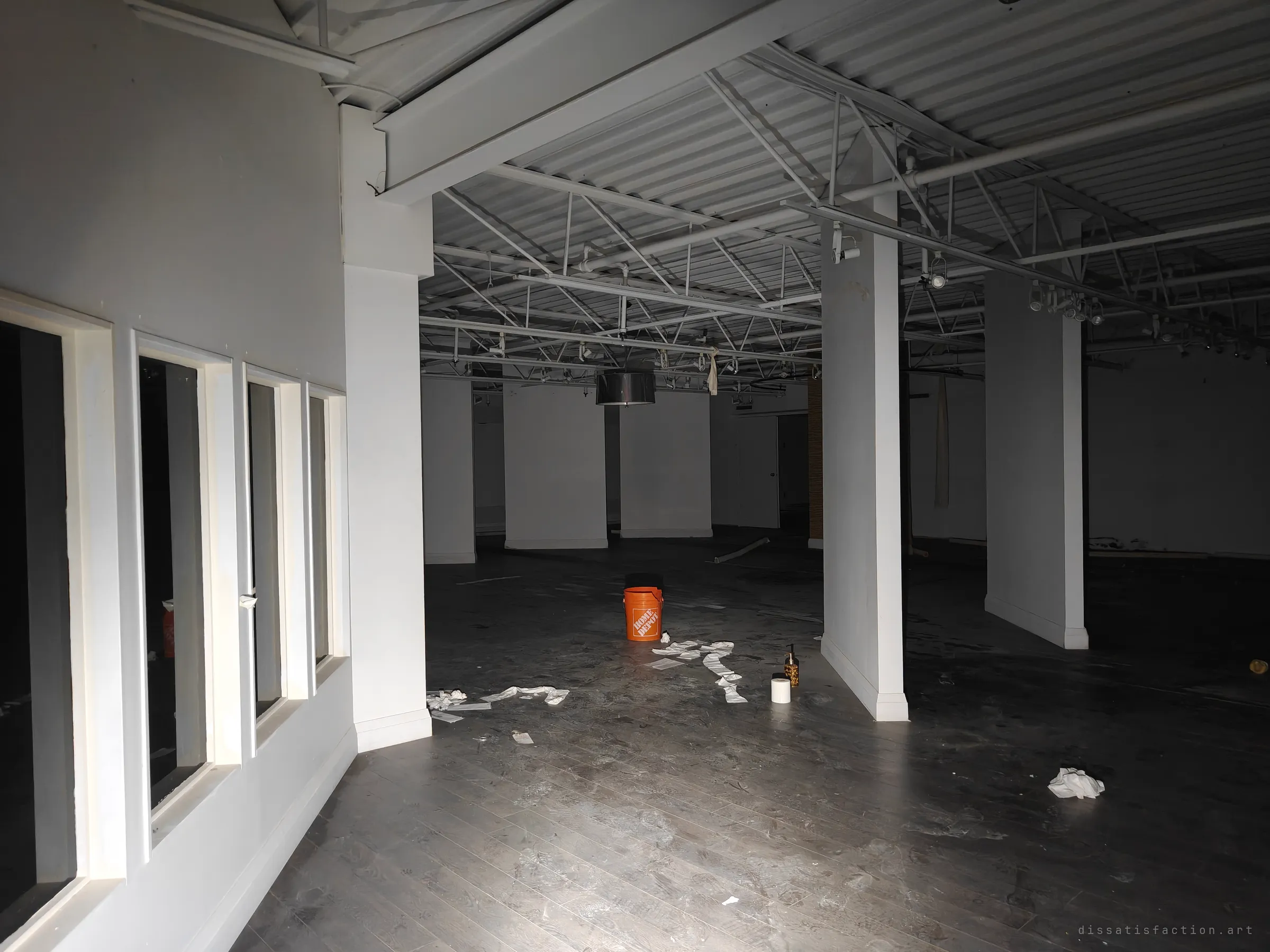 A long, dark showroom floor broken up by white pillars and exposed ceiling trusses, an orange bucket and scattered paper in the middle. The torchlight fades into the back of the unit.