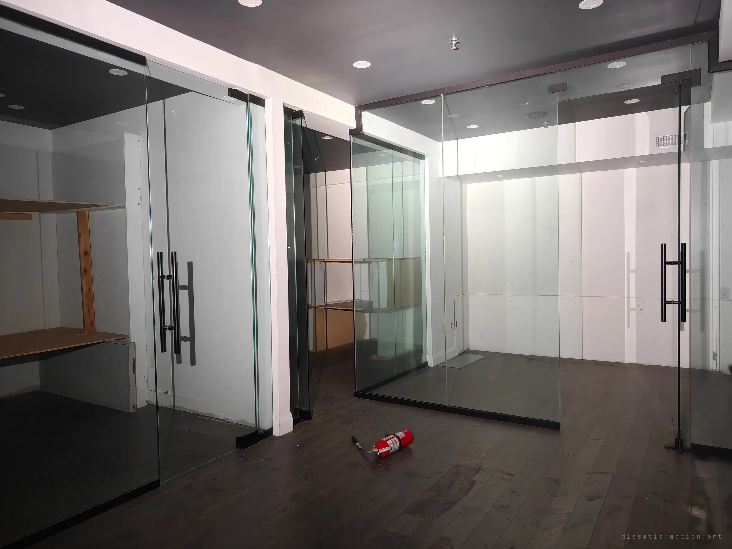 Frameless glass offices with black bar handles stand empty, a fire extinguisher lying on the wooden floor between them. Bare shelving is all that is left inside.